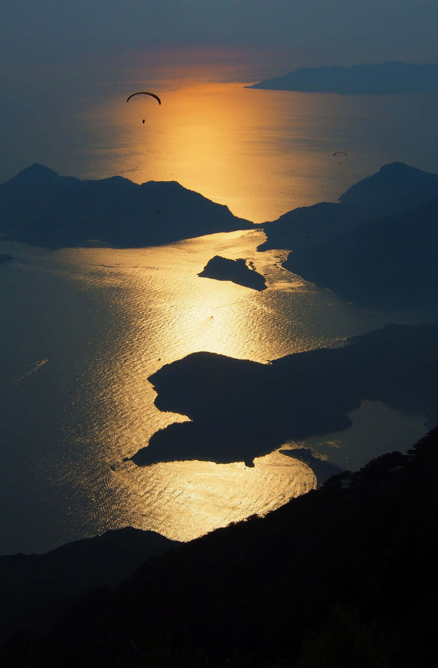 1488x2266 桌布 壁紙 — 夕陽在蜿蜒的海灣上閃耀著金色光芒，滑翔傘在剪影般的島嶼和山丘上空翱翔。