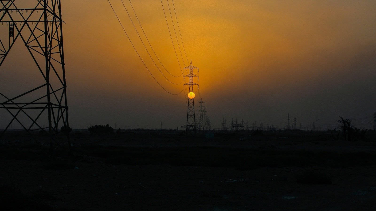 Dim 桌布 壁紙 — 夕陽在輸電塔後發光，在黑暗的工業景觀上投下長長的影子。