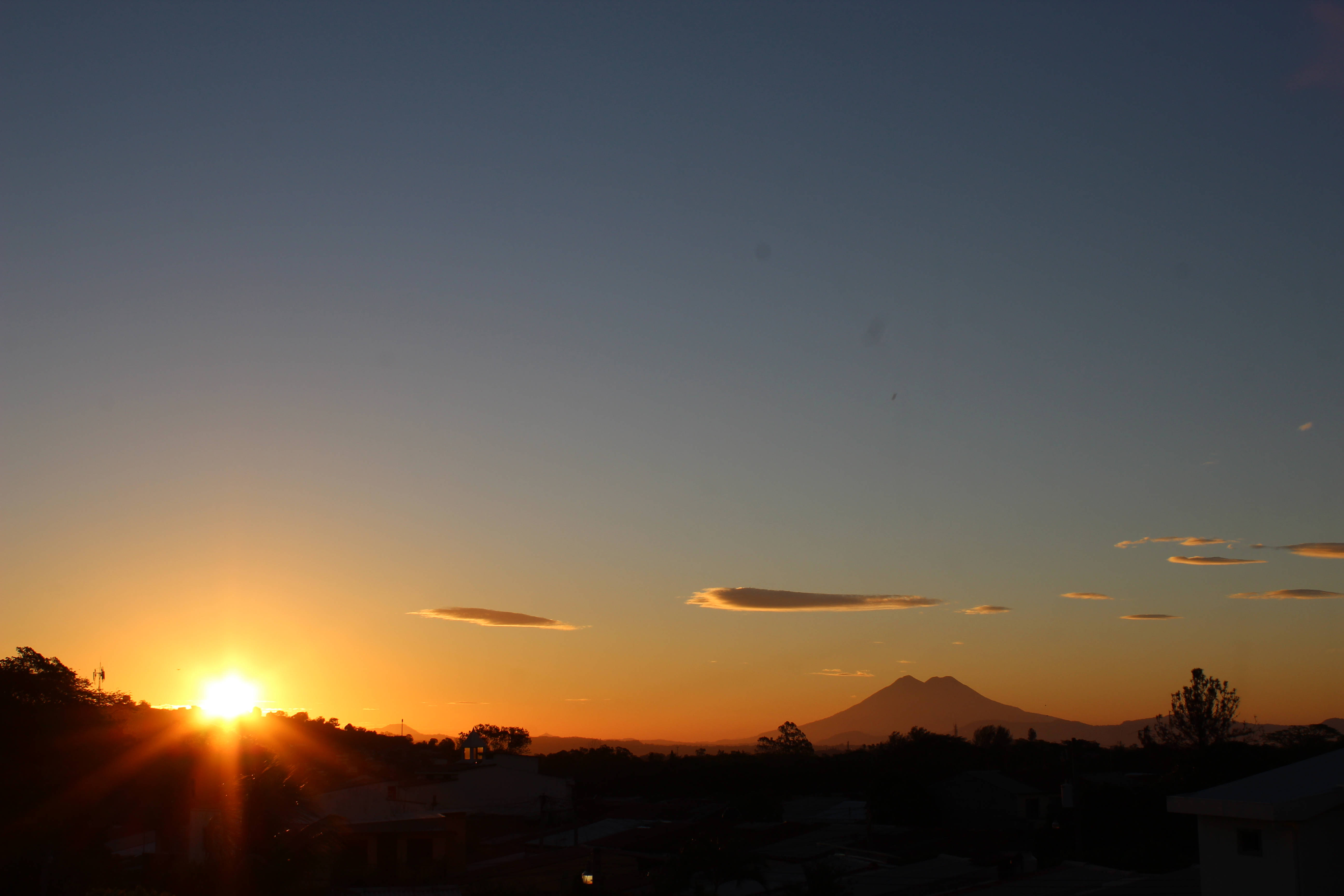 El Salvador 桌布 壁紙 — 夕陽在遠山後方閃耀，剪影般的屋頂和樹木勾勒出地平線。