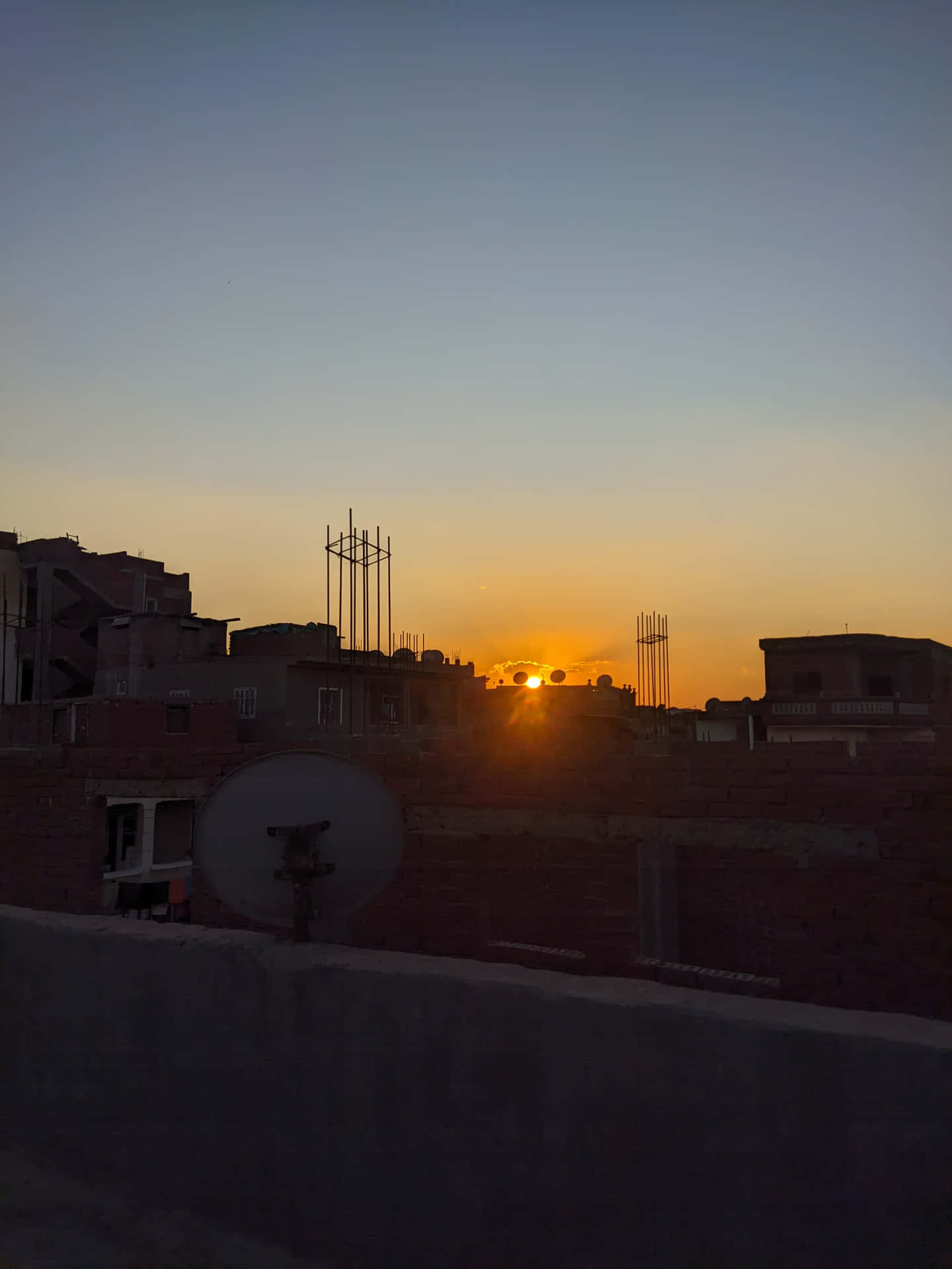 wallpaper 壁紙 — 夕陽在都市屋頂後方發出橙色光芒，衛星天線和未完工的建築物在漸變的天空中形成剪影。