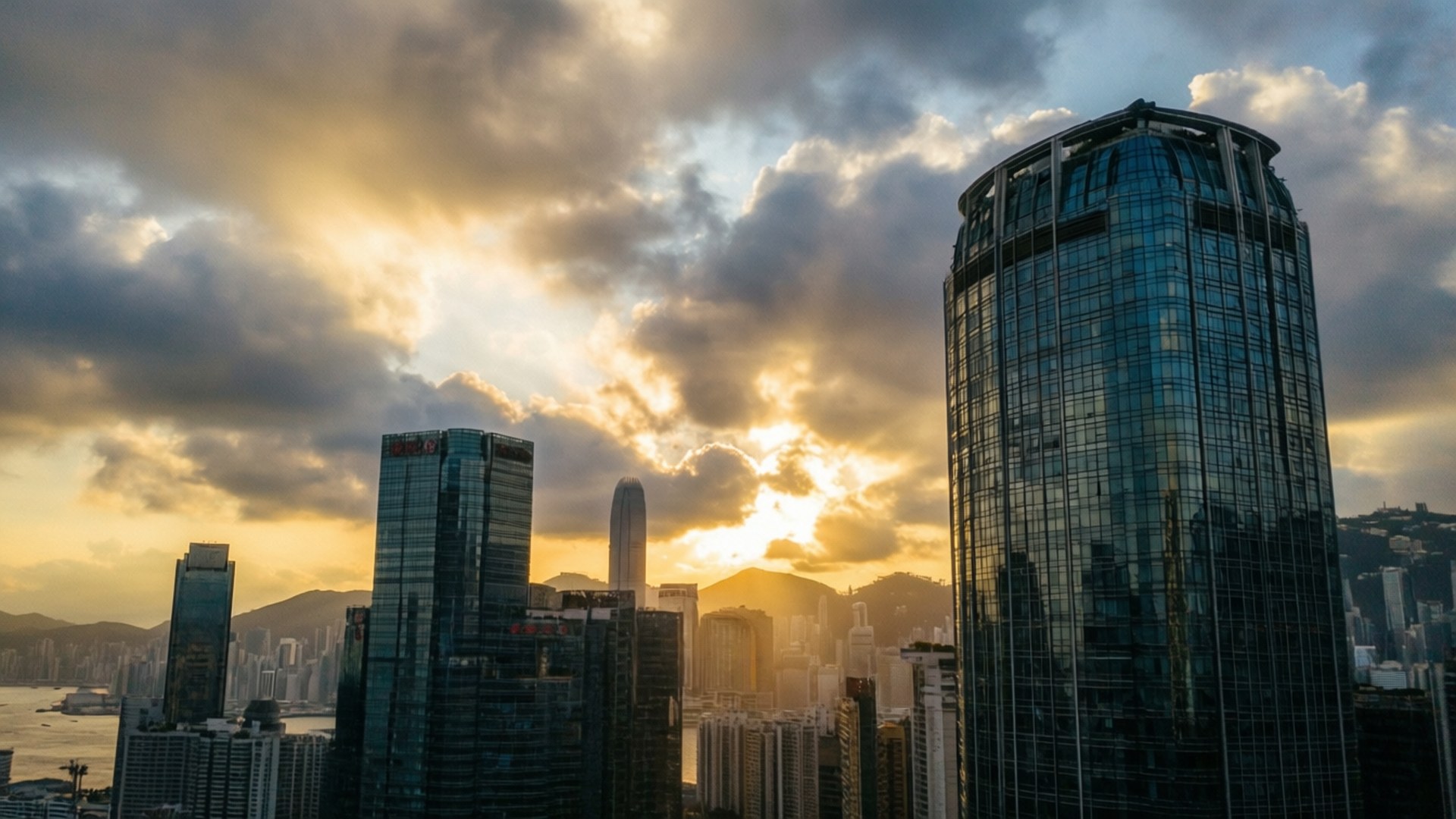 夕陽塔樓全景 壁紙 — 夕陽穿過戲劇性的雲層，照耀在一群密集的玻璃摩天大樓上，反射出金色的光芒。