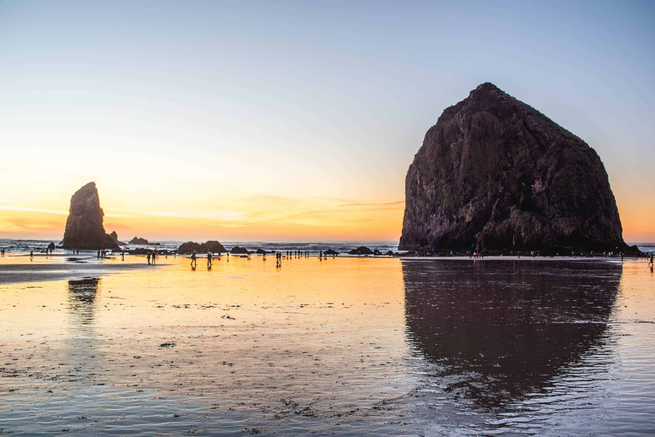 Oregon Coast 桌布 壁紙 — 夕陽將Haystack Rock和較小的海蝕柱沐浴在溫暖的金色光芒中，人們沿著濕漉漉的海灘漫步。
