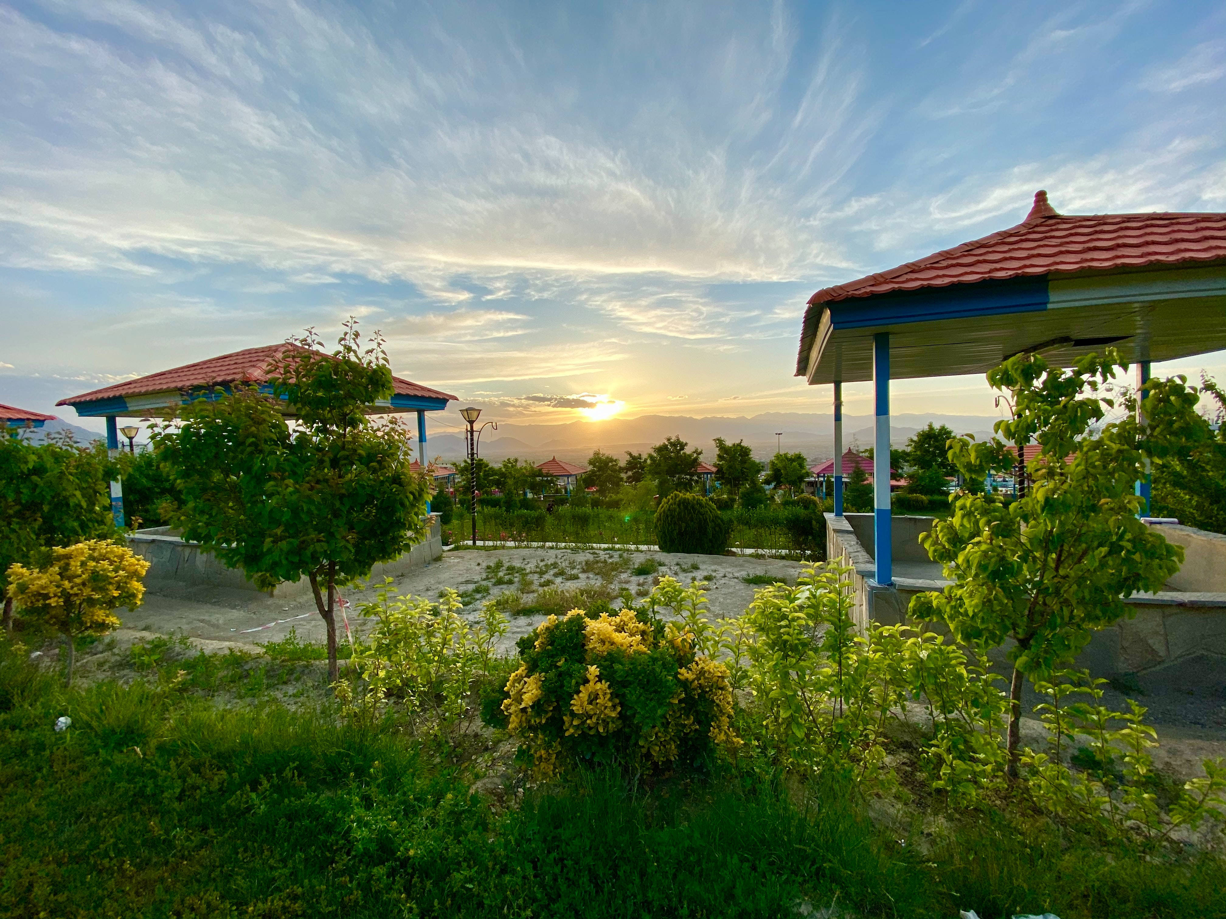 Kabul 桌布 壁紙 — 夕陽將一個寧靜的公園、紅頂涼亭和茂密綠植沐浴在溫暖的金色光芒中。
