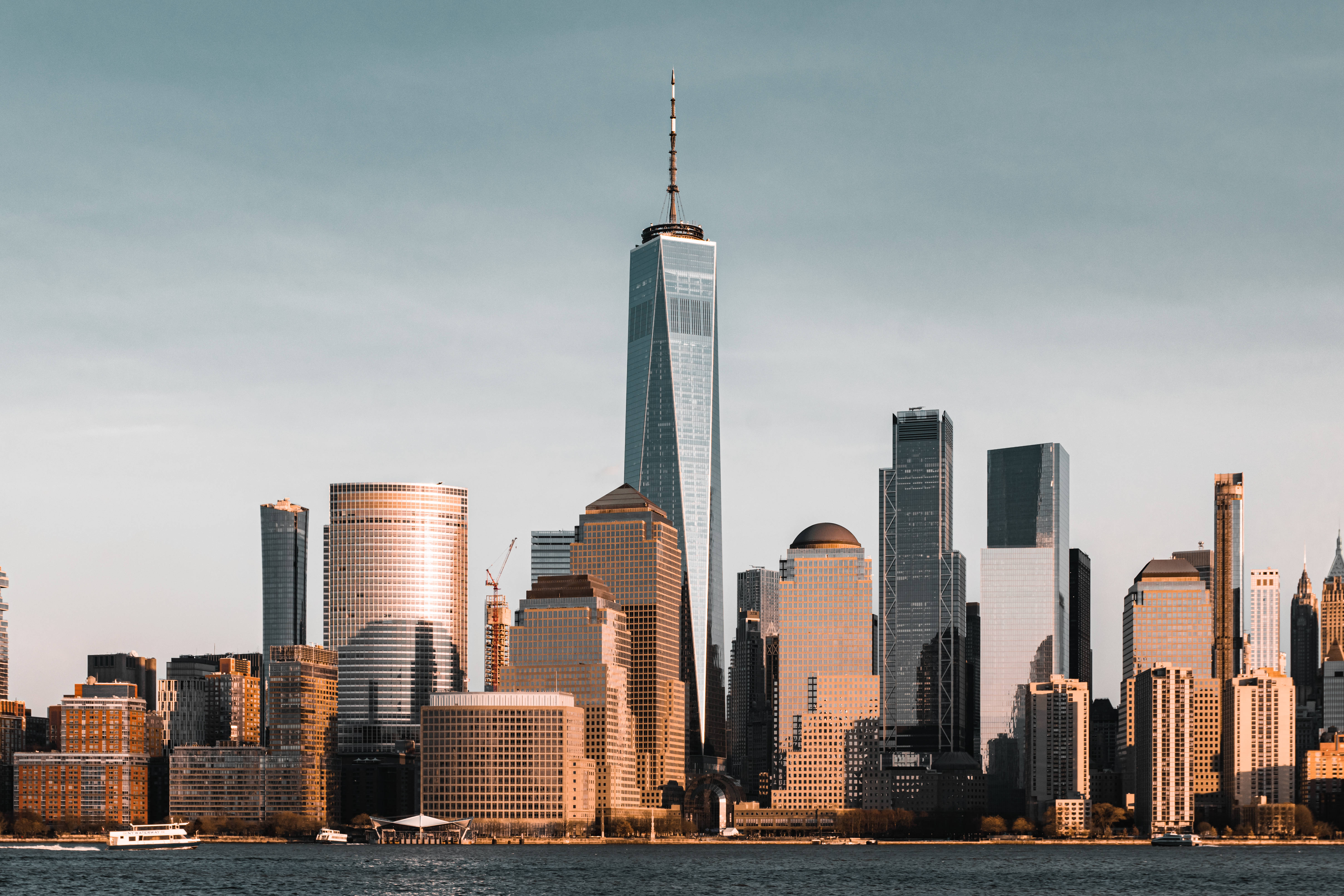 Nyc 桌布 壁紙 — 夕陽將世界貿易中心一號大樓和紐約市天際線沐浴在溫暖的金色光芒中，光線從玻璃塔樓和水面反射出來。