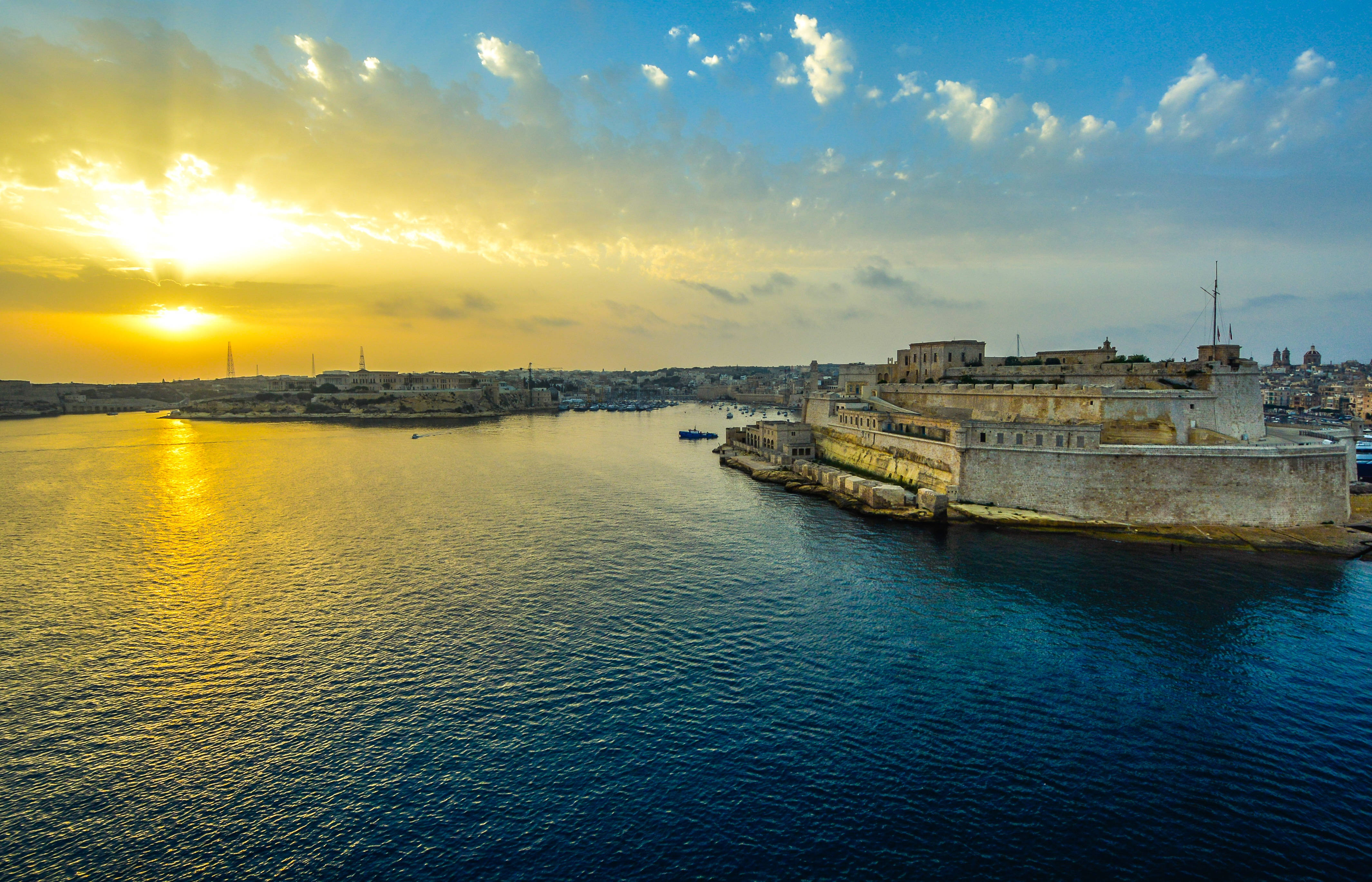 Malta 桌布 壁紙 — 夕陽將古老石堡和港口沐浴在金黃色光芒中，伴隨著漣漪的藍色海水和散落的雲朵。