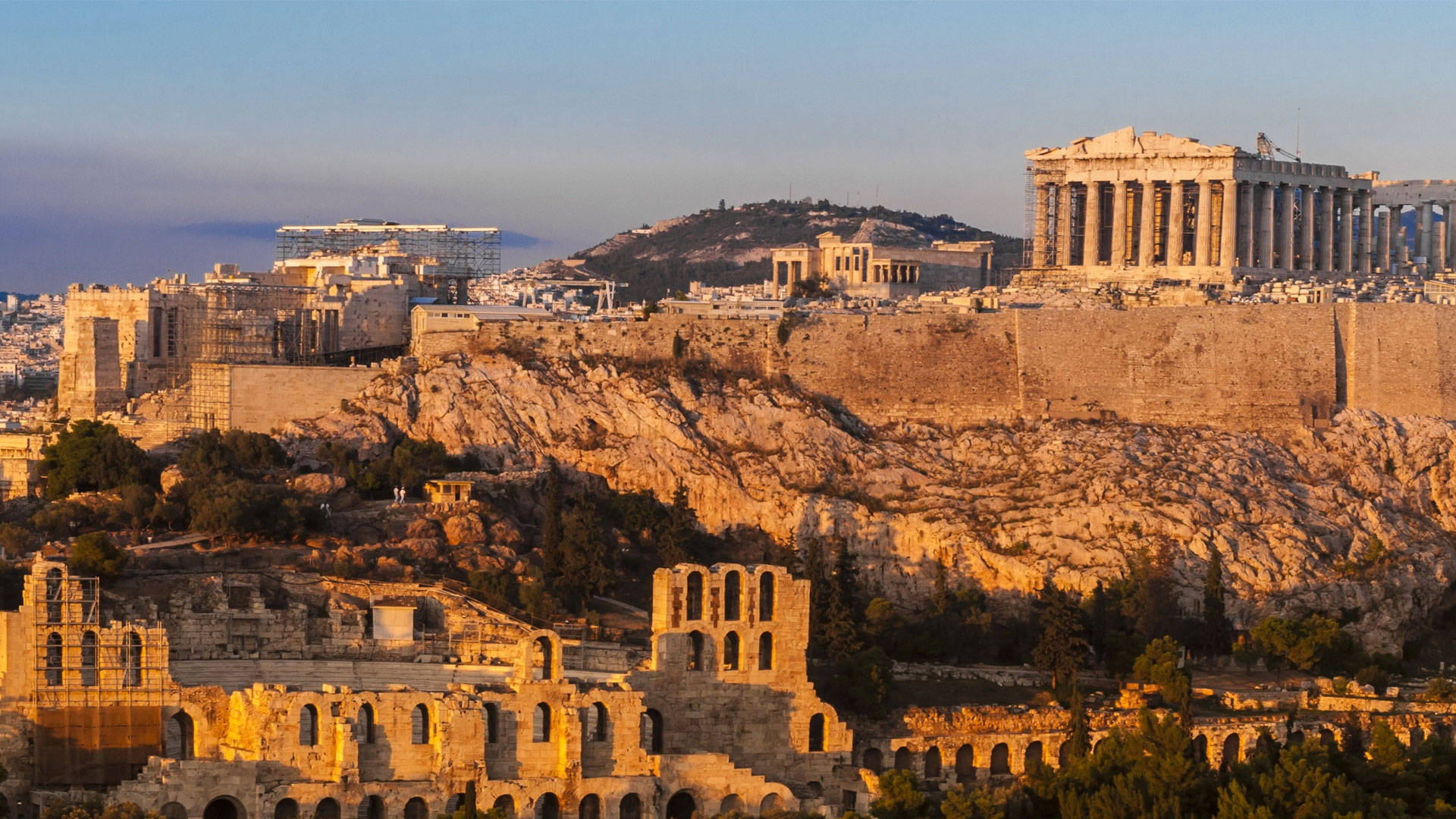 Athens 桌布 壁紙 — 夕陽將古雅典沐浴在金色光芒中，突顯帕德嫩神廟和廢墟劇院，映襯著清澈的藍天。