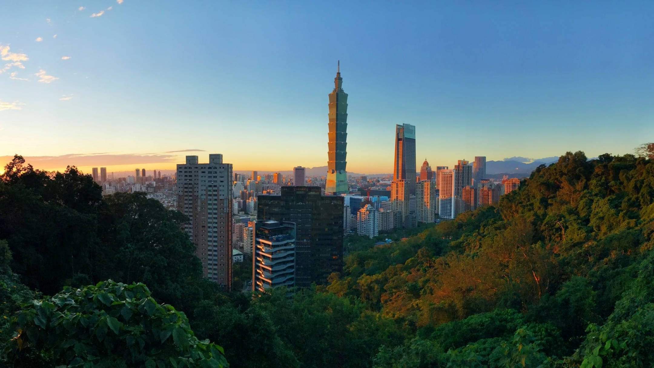 台北 桌布 壁紙 — 夕陽將台北的天際線沐浴在金色光芒中，鬱鬱蔥蔥的綠色山丘框住了高聳的城市景觀。