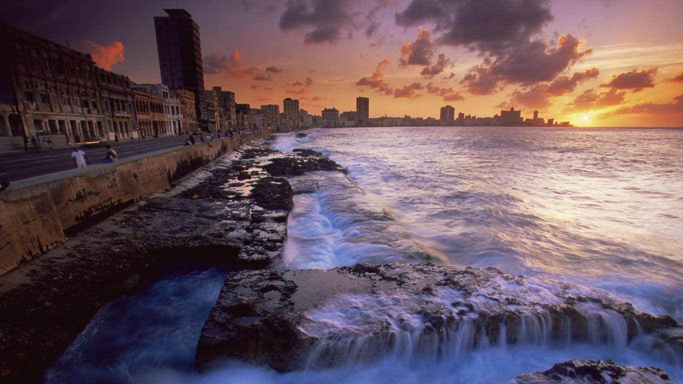夕陽天空 桌布 壁紙 — 夕陽將哈瓦那的天際線染成火熱的橙色，海浪拍打著歷史建築旁的岩石海岸。