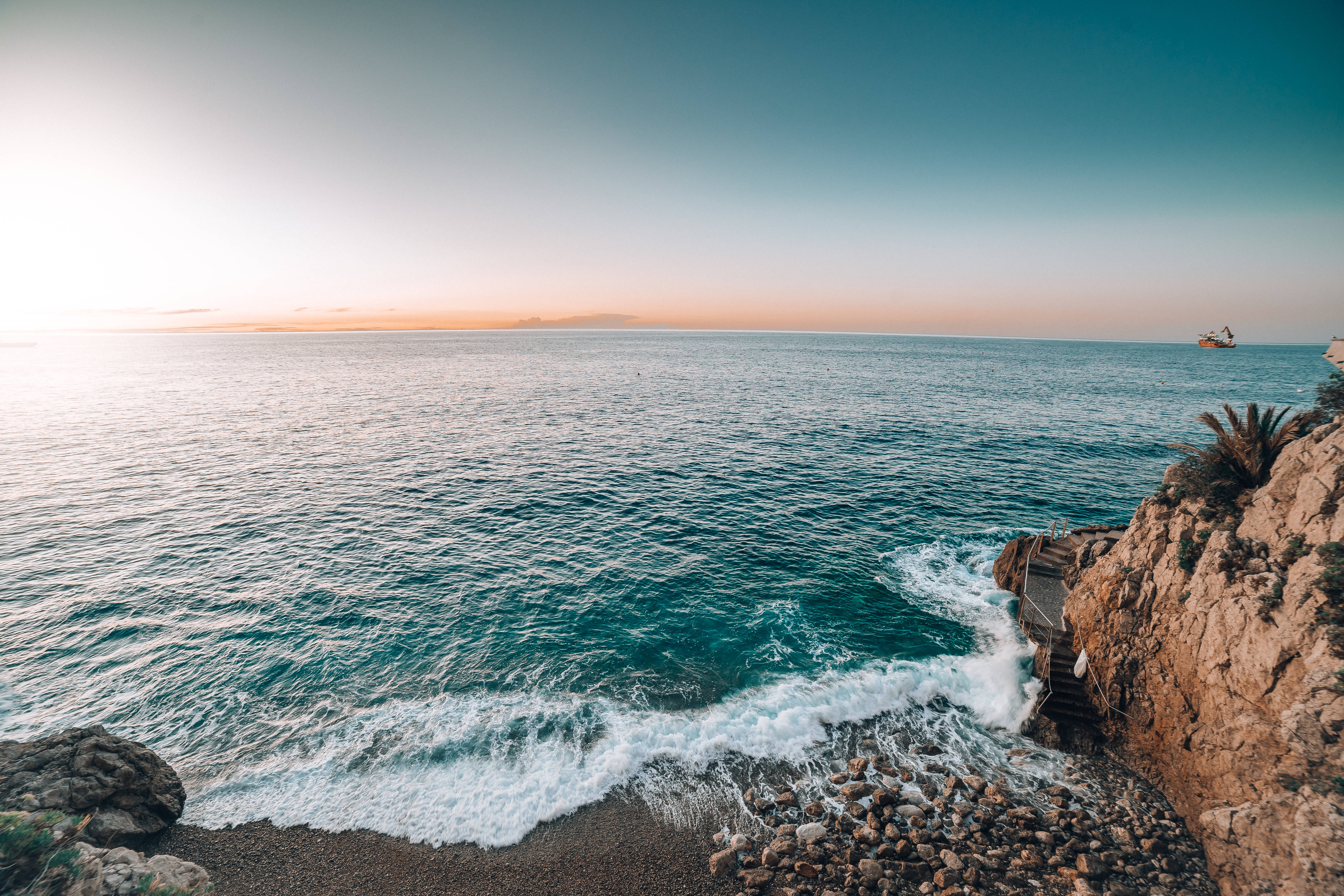 水 桌布 壁紙 — 夕陽將地平線染成橙色，海浪拍打岩石峭壁，遠處有一艘船在平靜的碧綠海面上。
