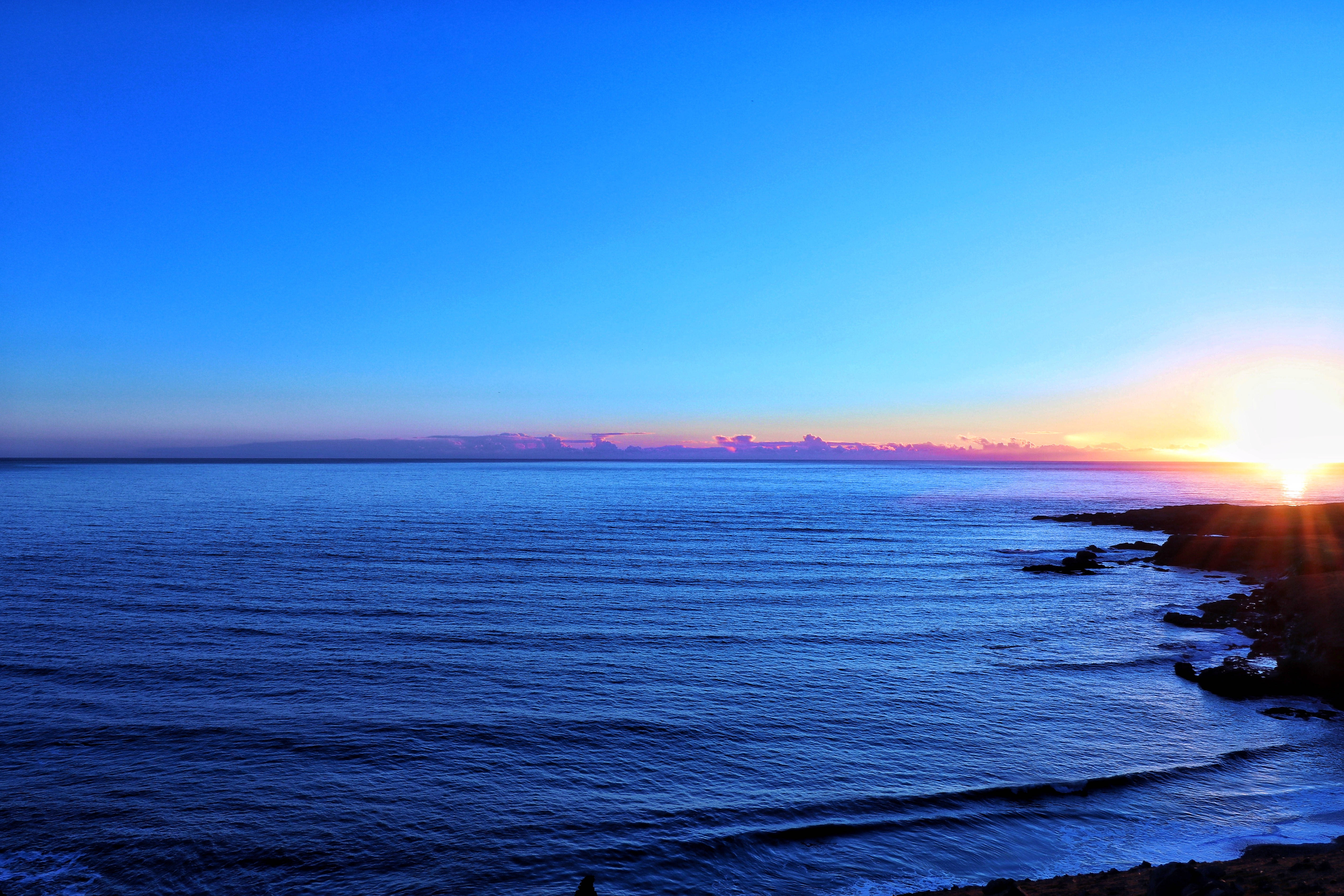 海洋 桌布 壁紙 — 夕陽將地平線染成火熱的橙色和粉紅色，沉入岩石海岸線之下，映襯著深藍色的海洋。