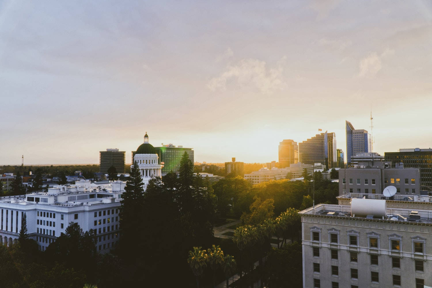 California 桌布 壁紙 — 夕陽將城市景觀沐浴在金色光芒中，摩天大樓和圓頂國會大廈聳立在綠樹成蔭的街道之上。