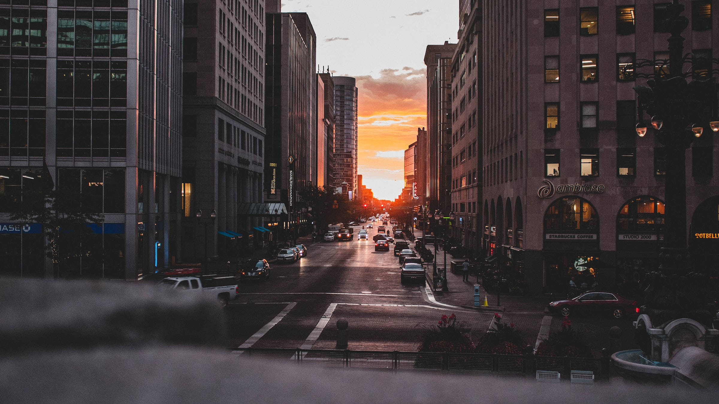 Buildings 桌布 壁紙 — 夕陽將城市街道染成溫暖的橙色，汽車在高聳的玻璃和石頭建築之間穿梭。