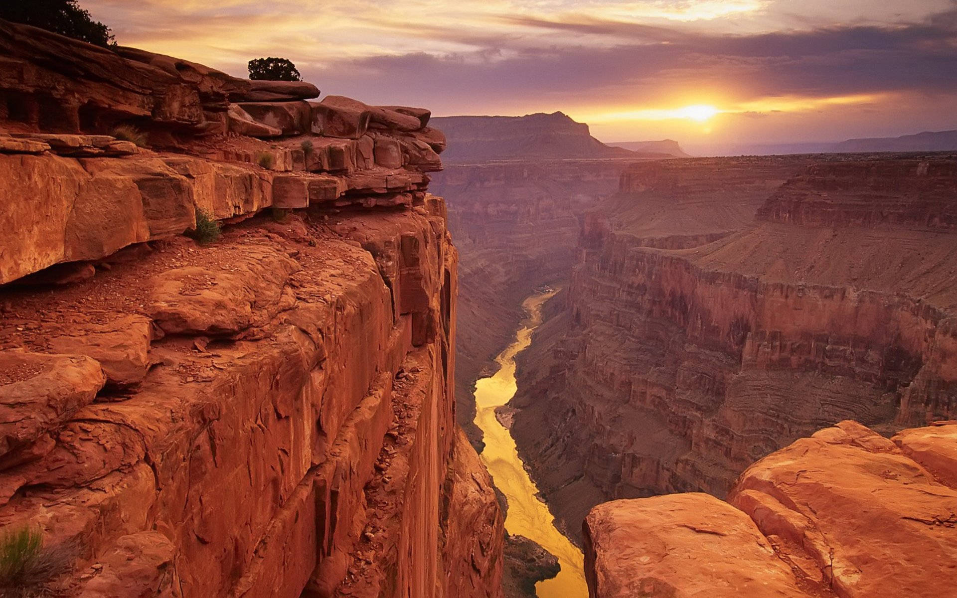 Arizona 桌布 壁紙 — 夕陽將大峽谷沐浴在溫暖的橙色光芒中，蜿蜒的河流在崎嶇的紅色岩石懸崖下閃爍。