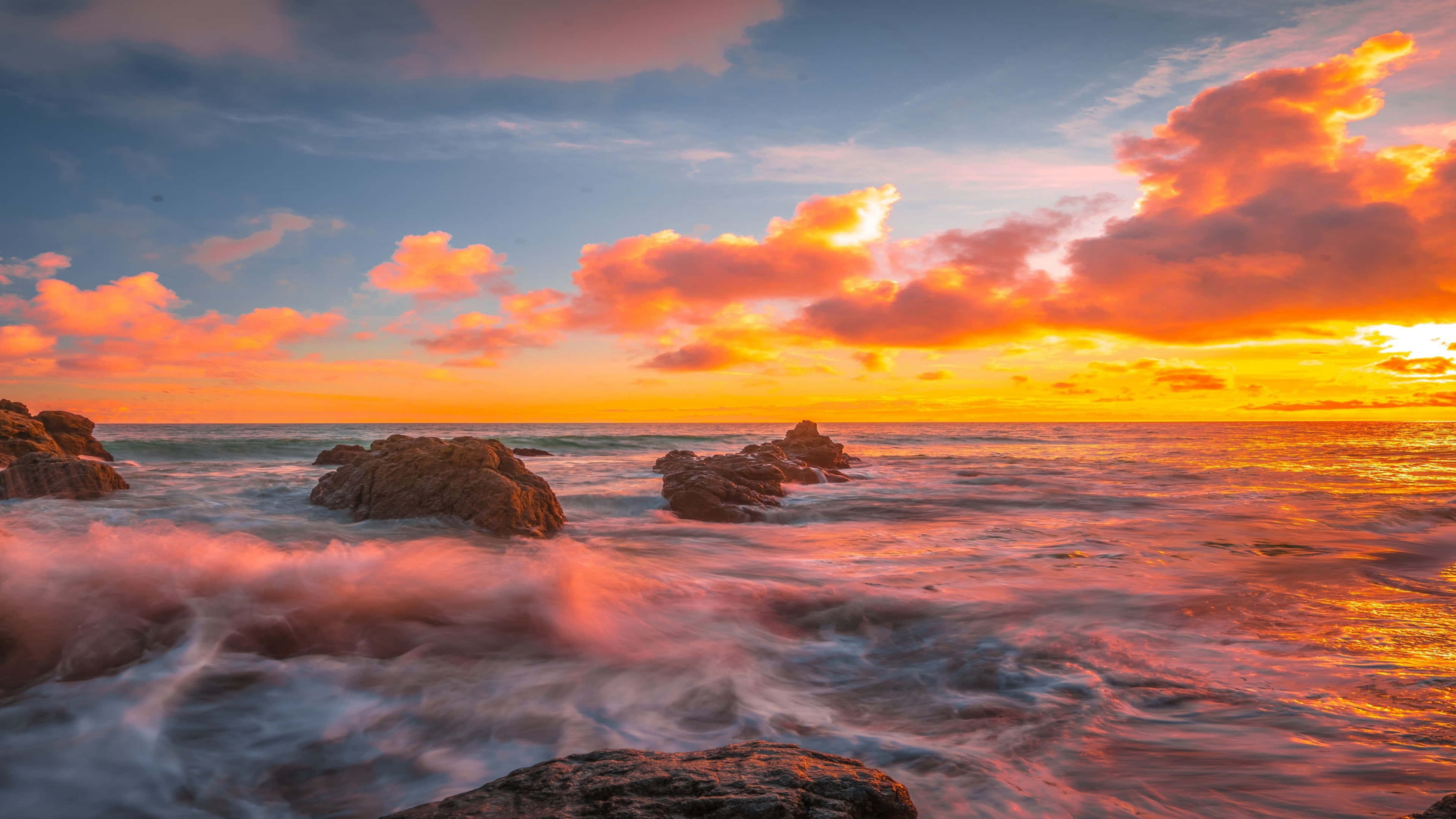 海景 桌布 壁紙 — 夕陽將天空和海洋染成火熱的橙色和粉紅色，海浪拍打著崎嶇的海岸岩石。