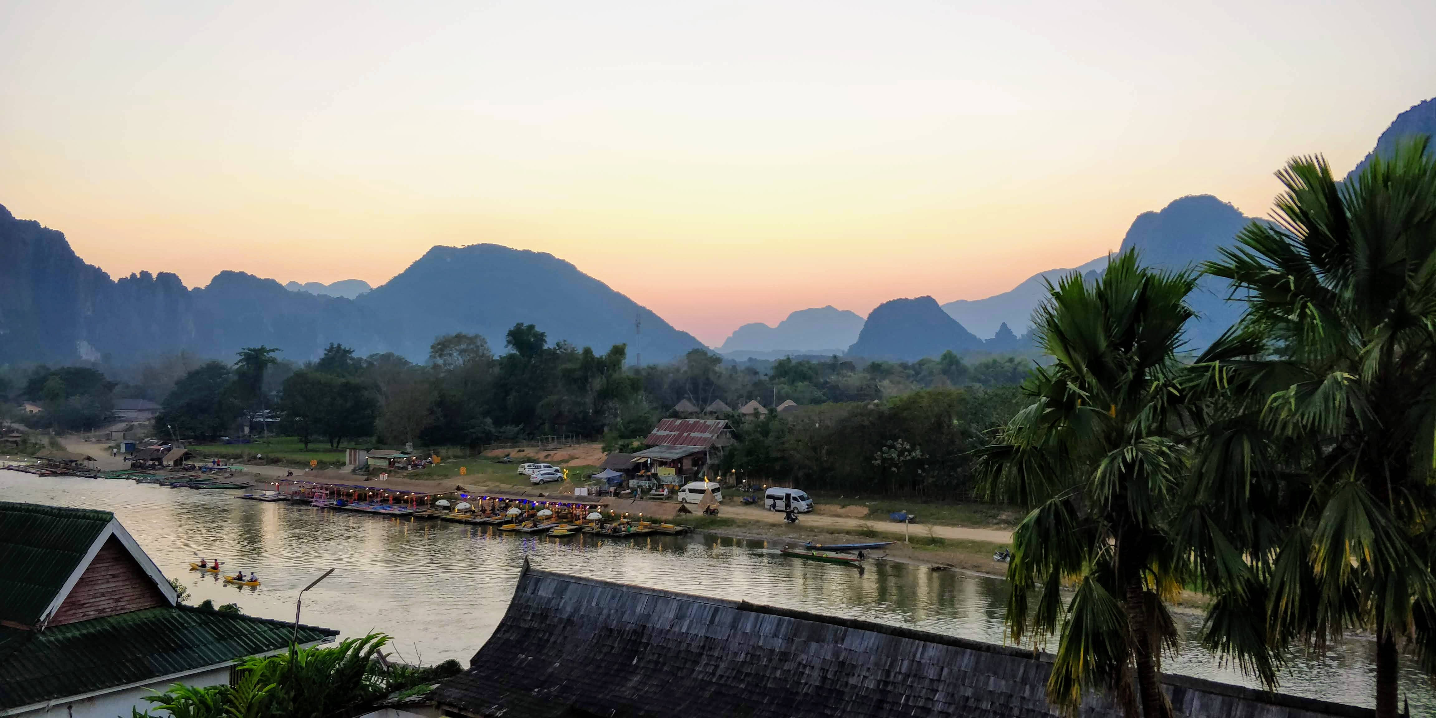 Laos 桌布 壁紙 — 夕陽將天空染成桃色和金色，映照著喀斯特山脈中棕櫚樹和皮划艇愛好者的河邊村莊。