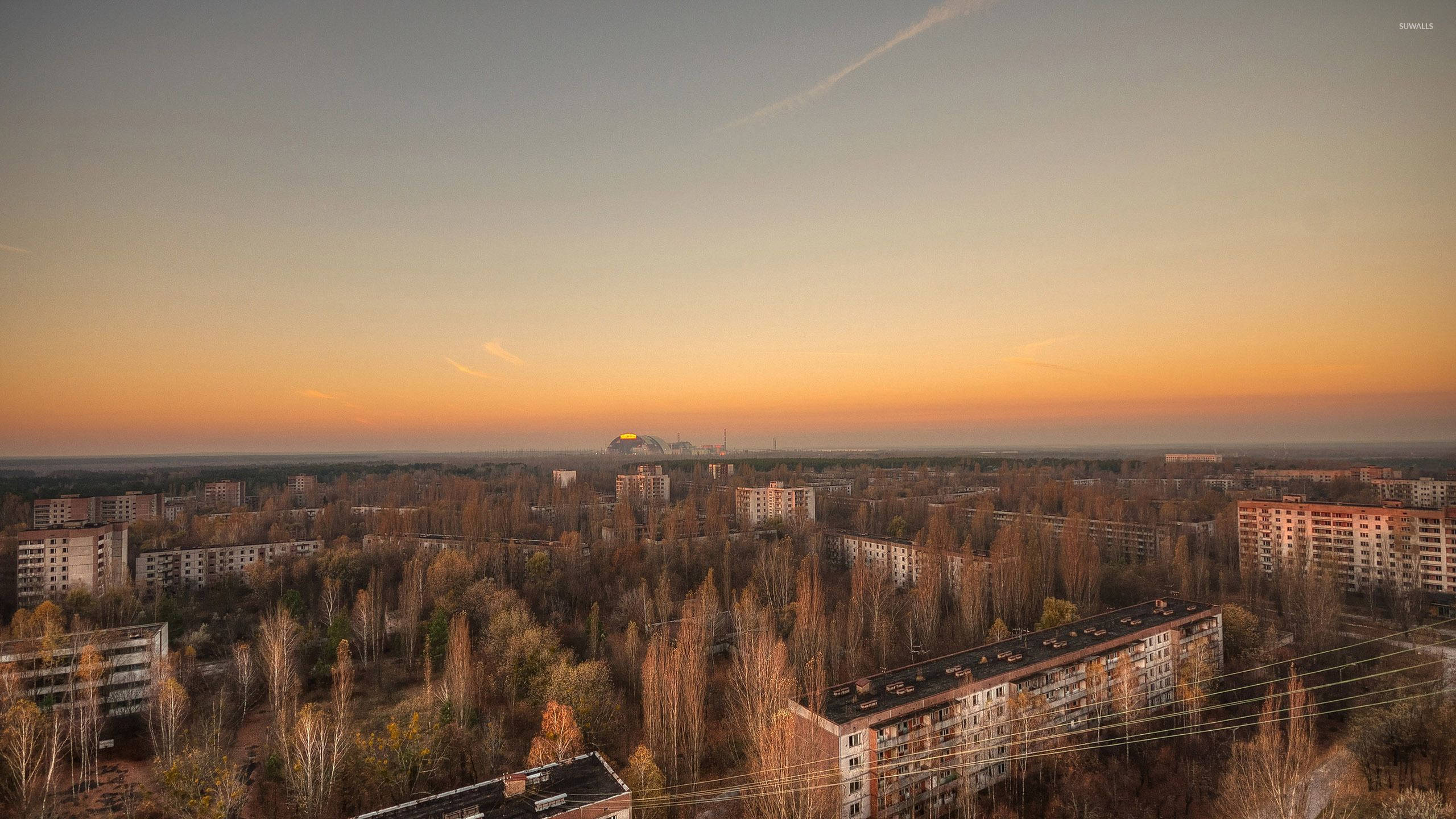 風景 桌布 壁紙 — 夕陽將天空染成橘色，映照著被遺棄的蘇聯時代公寓大樓和無葉的樹木，形成一片寂靜的城市荒地。