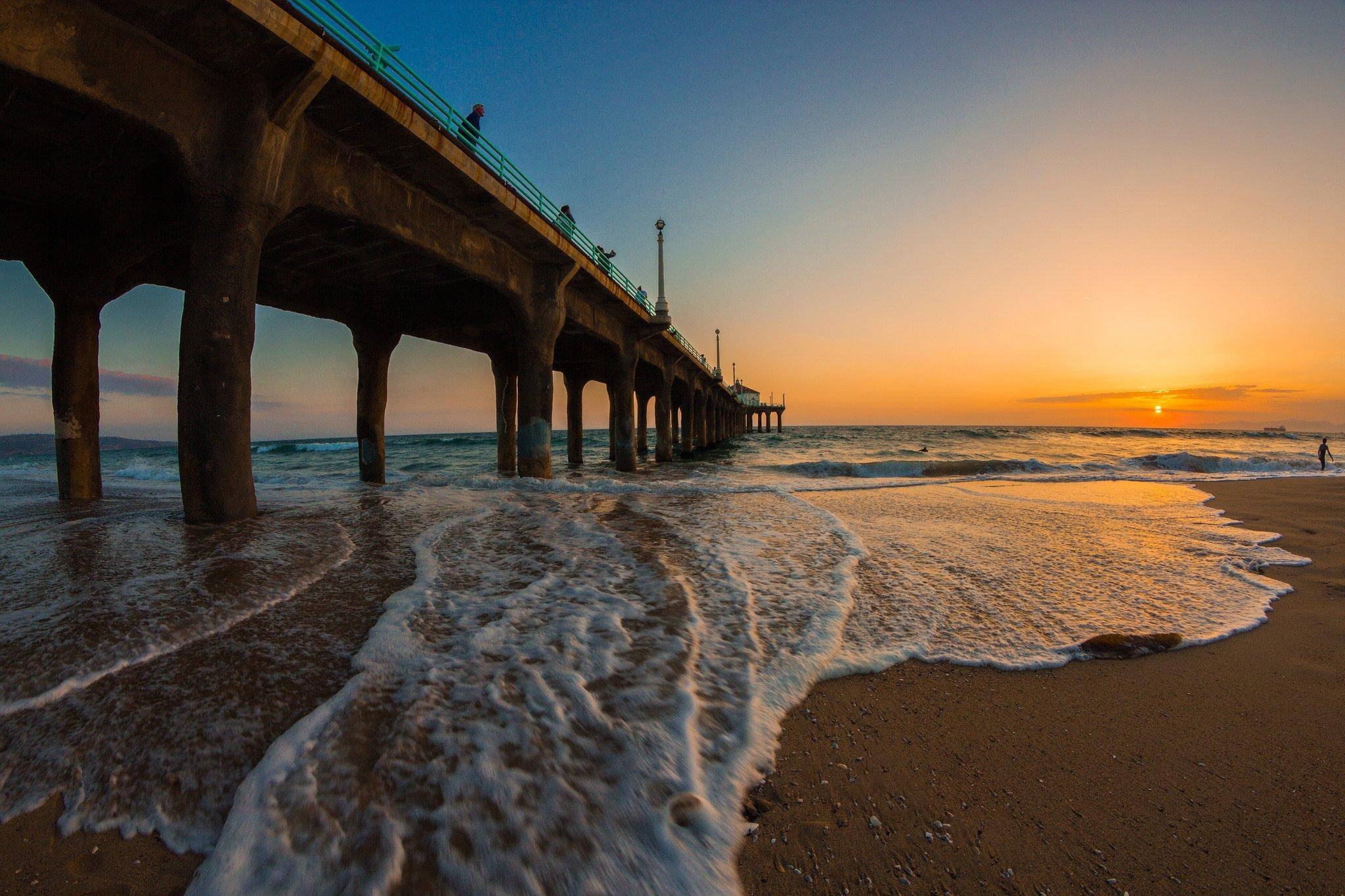 Venice Beach 桌布 壁紙 — 夕陽將天空染成橙色，海浪在長碼頭下拍打，人們沿著欄杆行走。