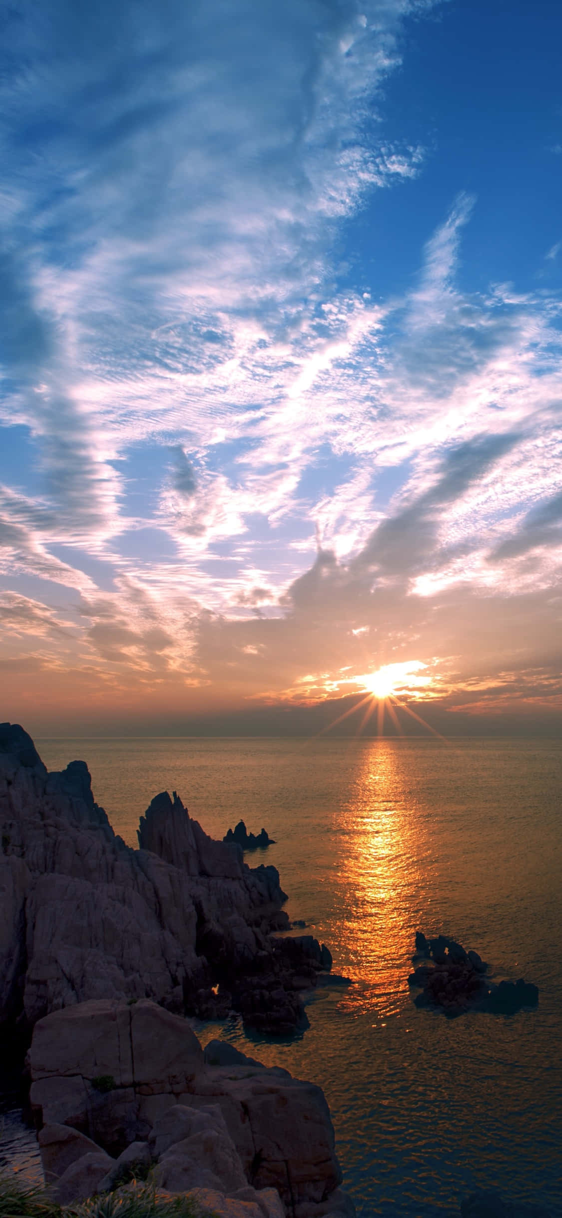 Sunrise iPhone 桌布 壁紙 — 夕陽將天空染成橙色和粉紅色，金色的光芒在平靜的海面上反射，旁邊是崎嶇的沿海岩石。