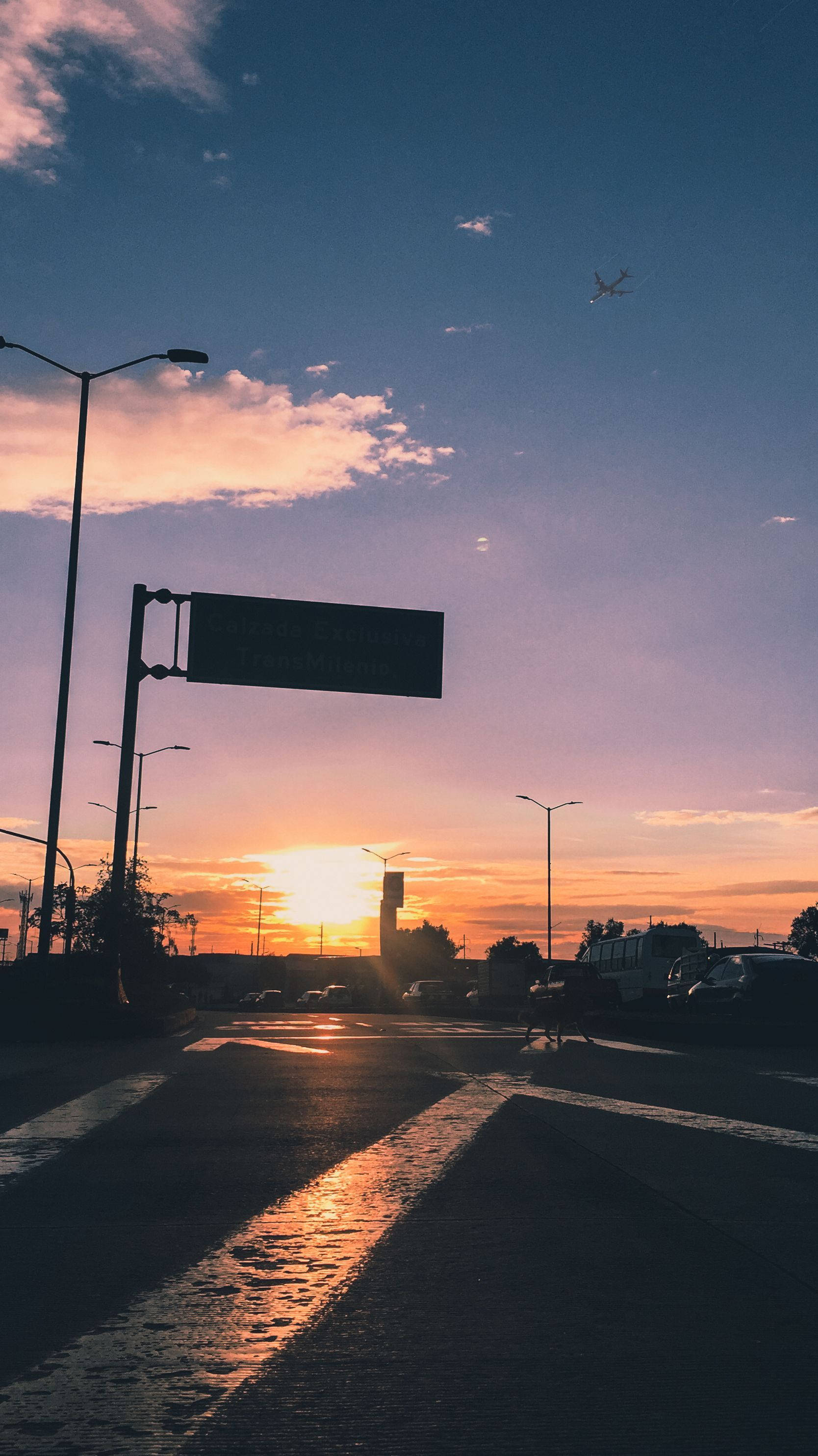 Bogota 桌布 壁紙 — 夕陽將天空染成橙色和紫色，一架孤獨的飛機飛過濕漉漉的城市街道，街燈和汽車形成剪影。