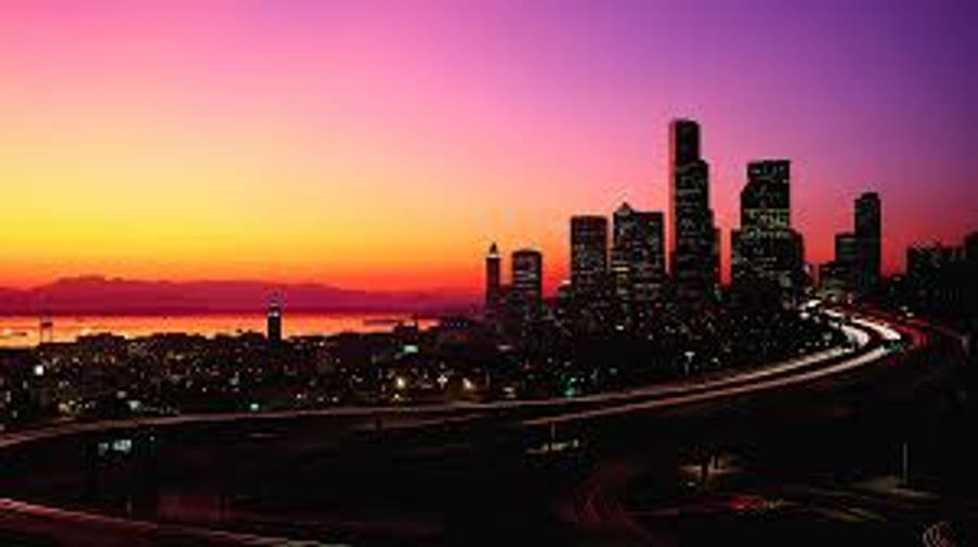 Seattle Skyline 桌布 壁紙 — 夕陽將天空染成橙色和紫色，城市摩天大樓和閃爍的高速公路在地平線上形成剪影。