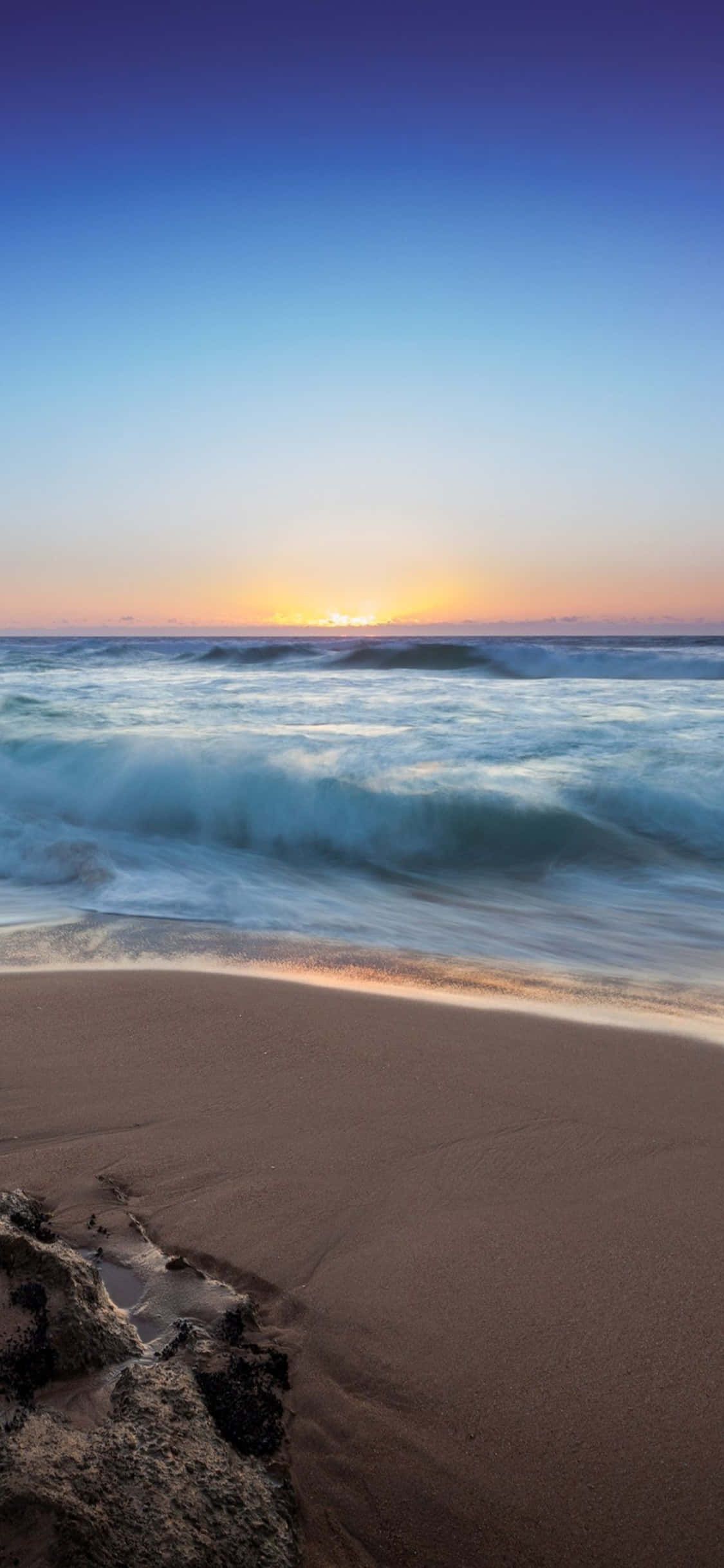 iPhone X Beach 桌布 壁紙 — 夕陽將天空染成橙色和紫色，海浪拍打沙灘，前景有深色岩石。