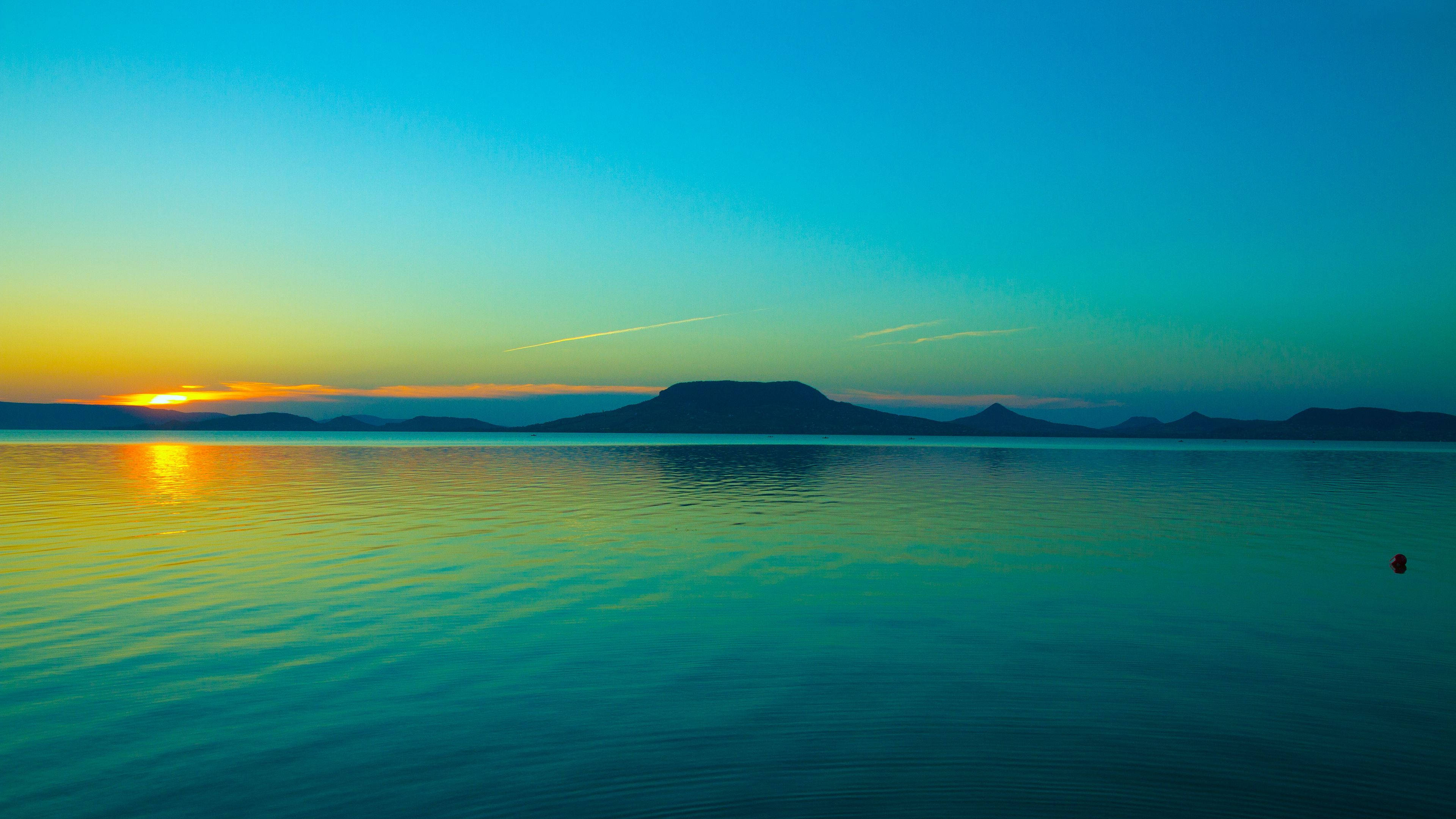 Ocean Sunset 桌布 壁紙 — 夕陽將天空染成溫暖的橙色和清涼的藍綠色，倒映在平靜的水面上，遠山輪廓依稀可見。