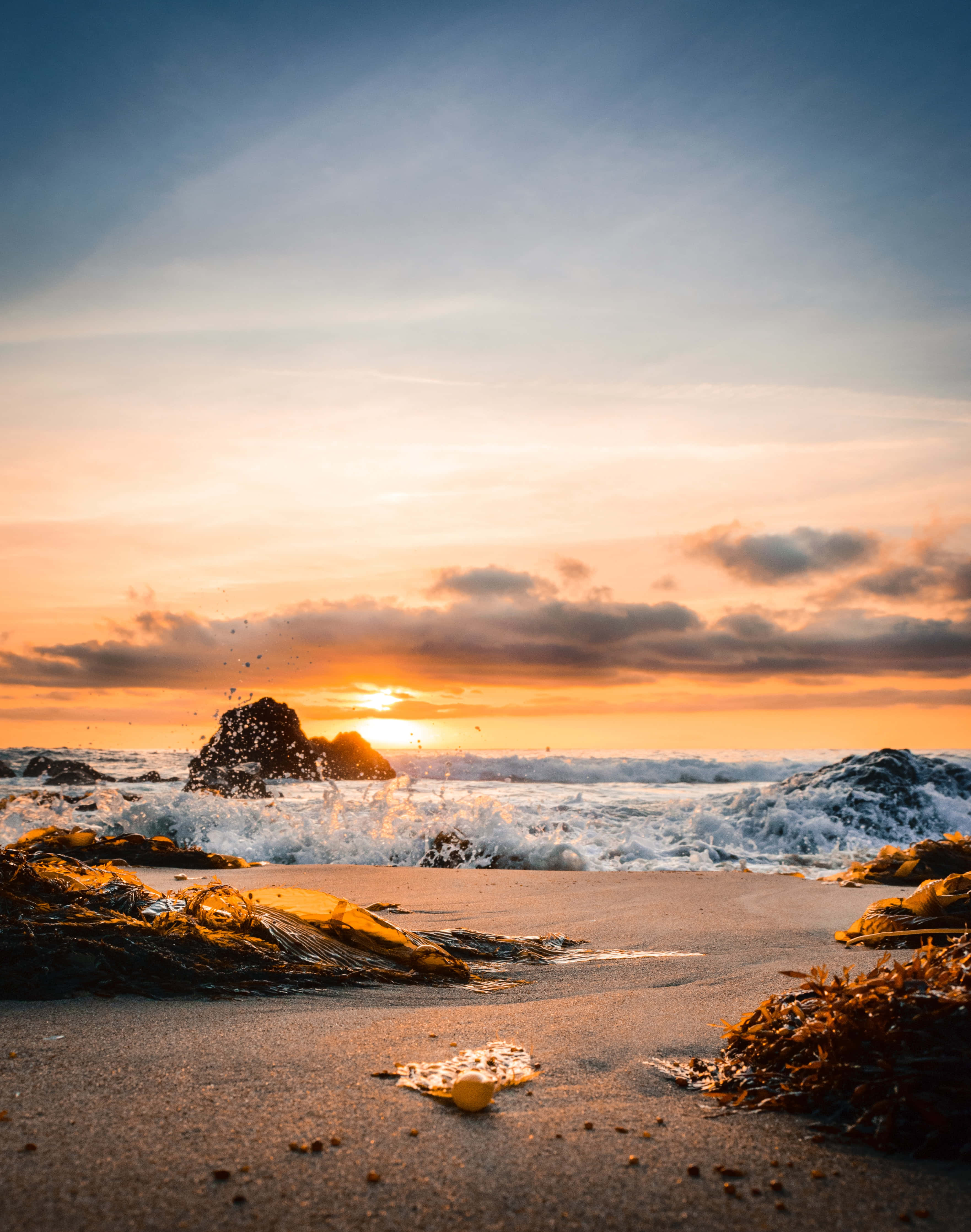 Sunset Beach 桌布 壁紙 — 夕陽將天空染成溫暖的橙色和粉紅色調，海浪拍打沙灘上的深色岩石。