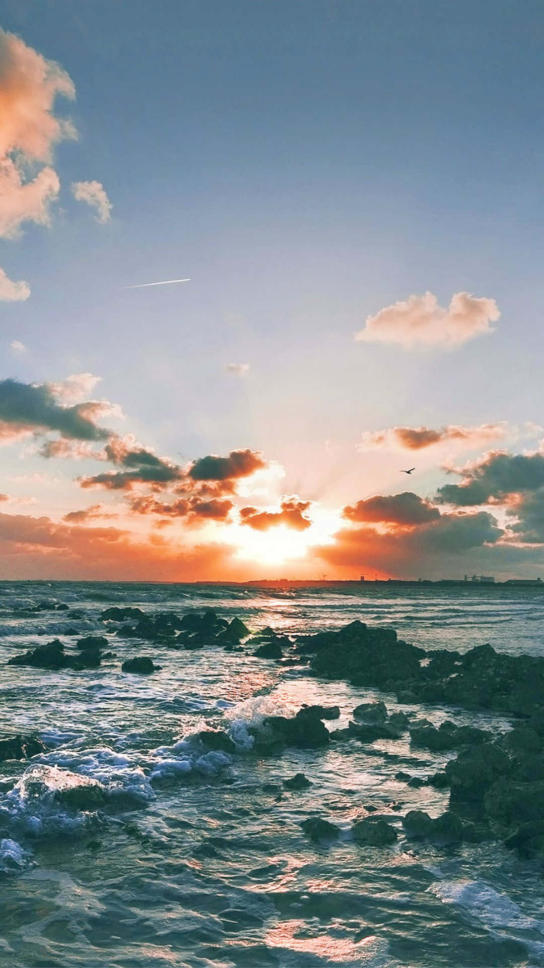 Sunset Beach iPhone 桌布 壁紙 — 夕陽將天空染成溫暖的橙色和粉紅色調，海浪拍打著海岸線上的黑色岩石。