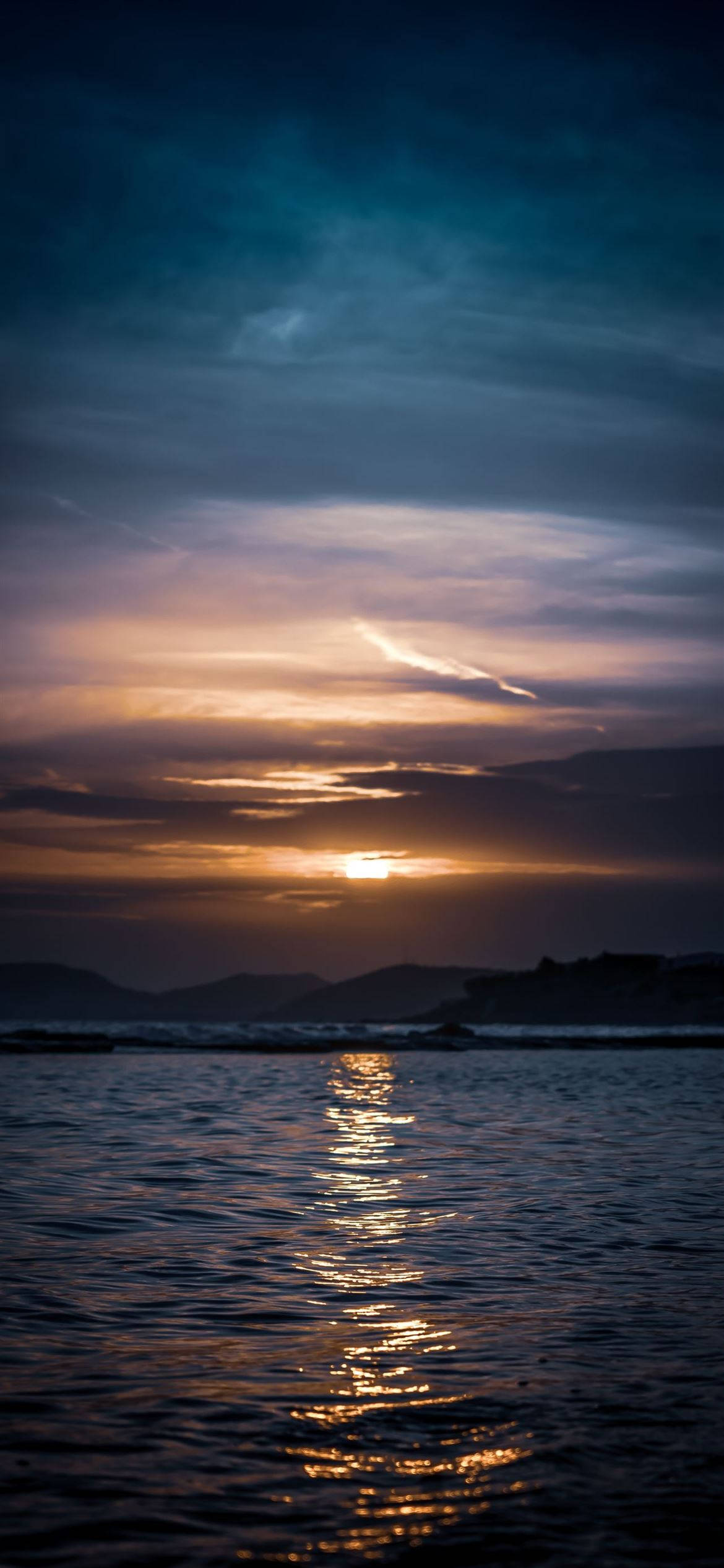 桌布 壁紙 — 夕陽將天空染成溫暖的金色和深藍色，它的倒影在遠處山丘附近平靜的海面上舞動。