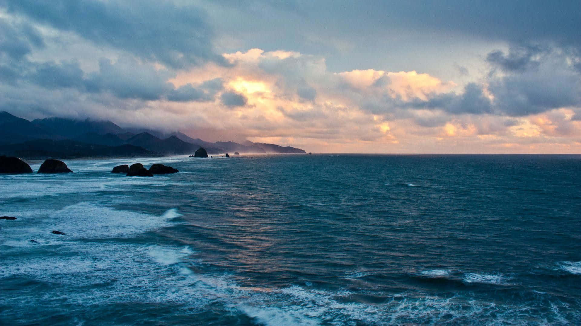 海灘 桌布 壁紙 — 夕陽將天空染成溫暖的金色和粉紅色，映照在波濤洶湧的海洋上，有深色岩石和遠山。
