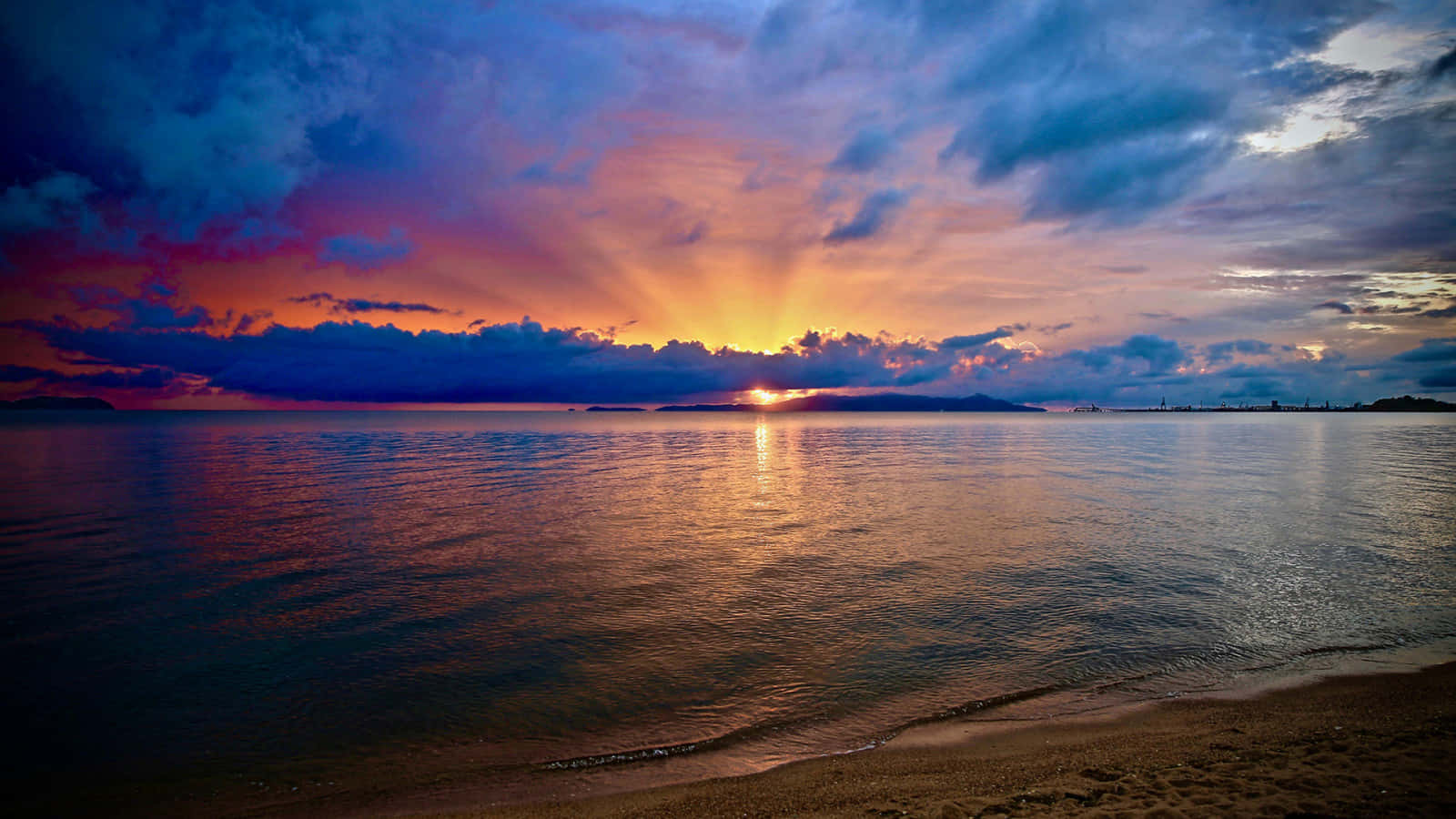 海灘日落 桌布 壁紙 — 夕陽將天空染成火橙色和深紫色，光線穿過雲層，映照在平靜的海面上，反射出金色的光芒。
