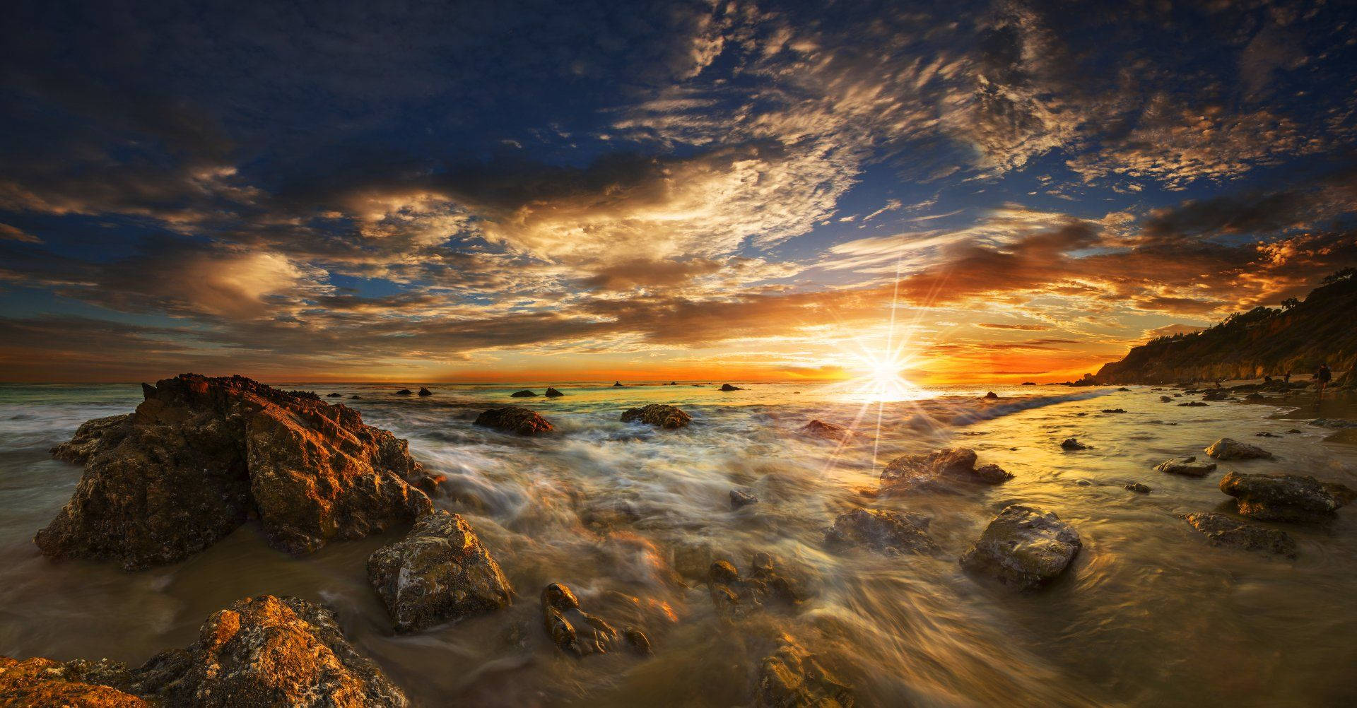 Malibu 桌布 壁紙 — 夕陽將天空染成火橙色和深藍色，海浪拍打崎嶇海岸線上的岩石。