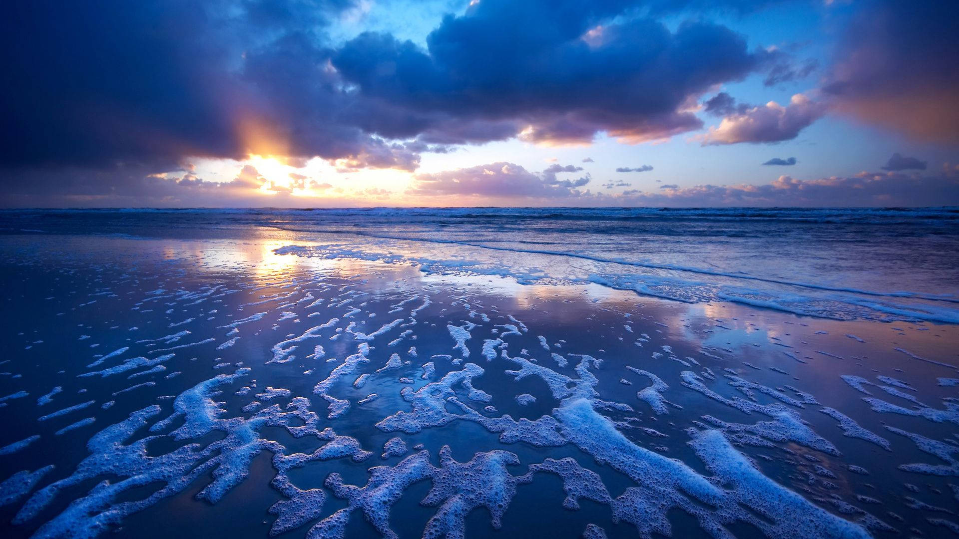 海邊夕照美景 壁紙 — 夕陽將天空染成火橙色和深藍色，海浪拍打濕沙灘，形成泡沫圖案。