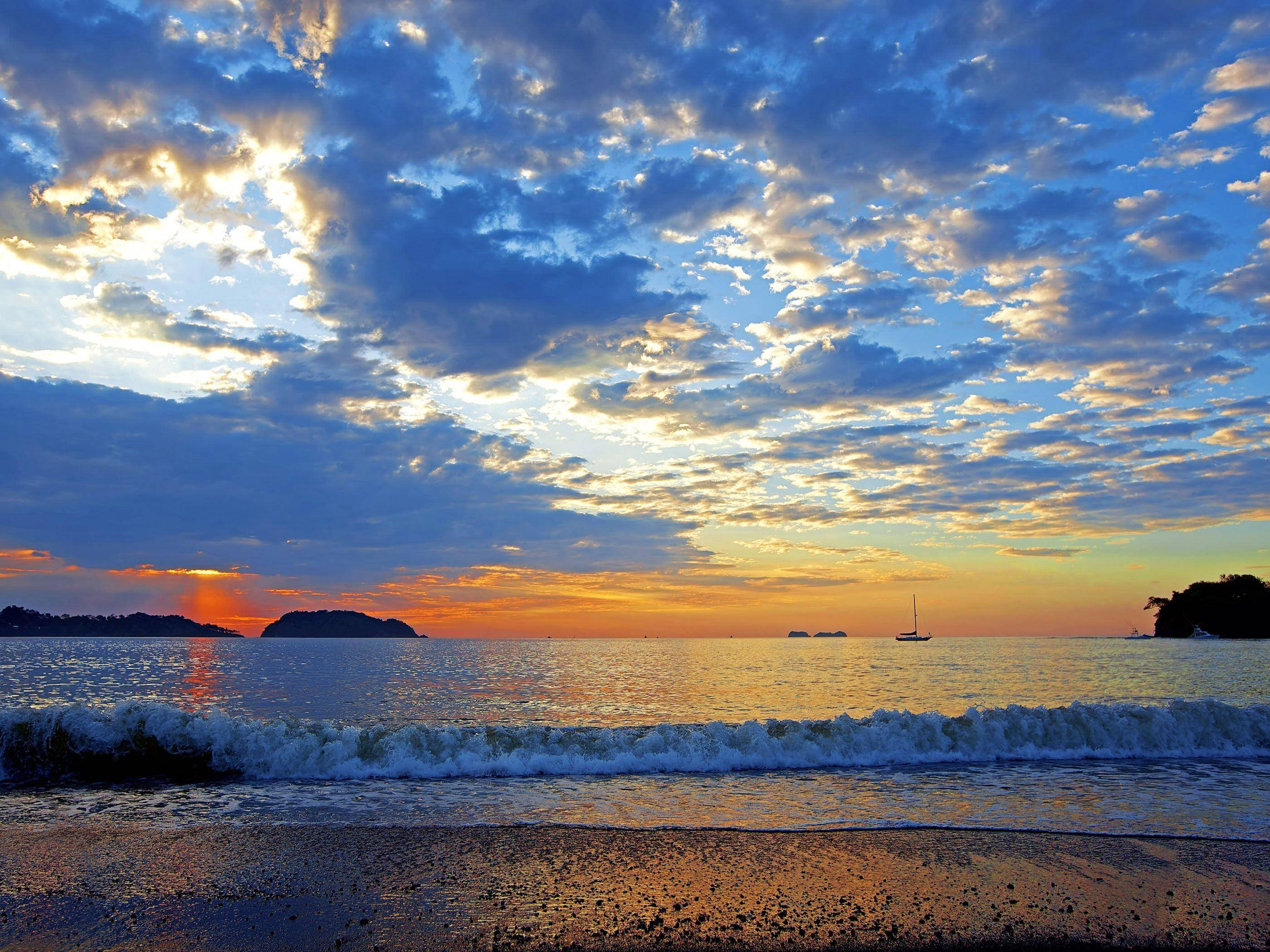 雲朵 桌布 壁紙 — 夕陽將天空染成火橙色和深藍色，海浪拍打沙灘，遠處帆船的剪影清晰可見。