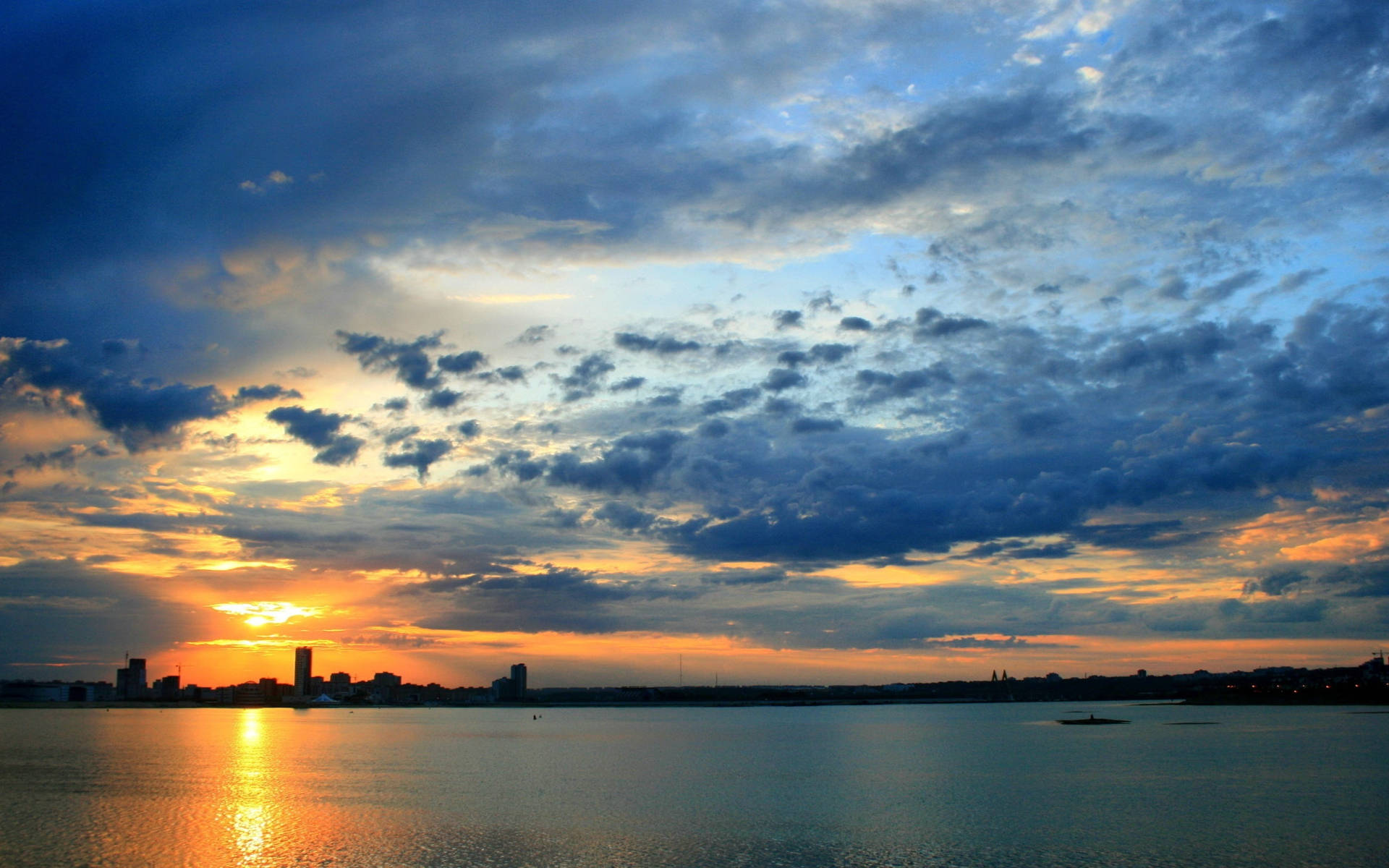 Kazan 桌布 壁紙 — 夕陽將天空染成火橙色和深藍色，雲朵倒映在平靜的水面上，城市天際線剪影。