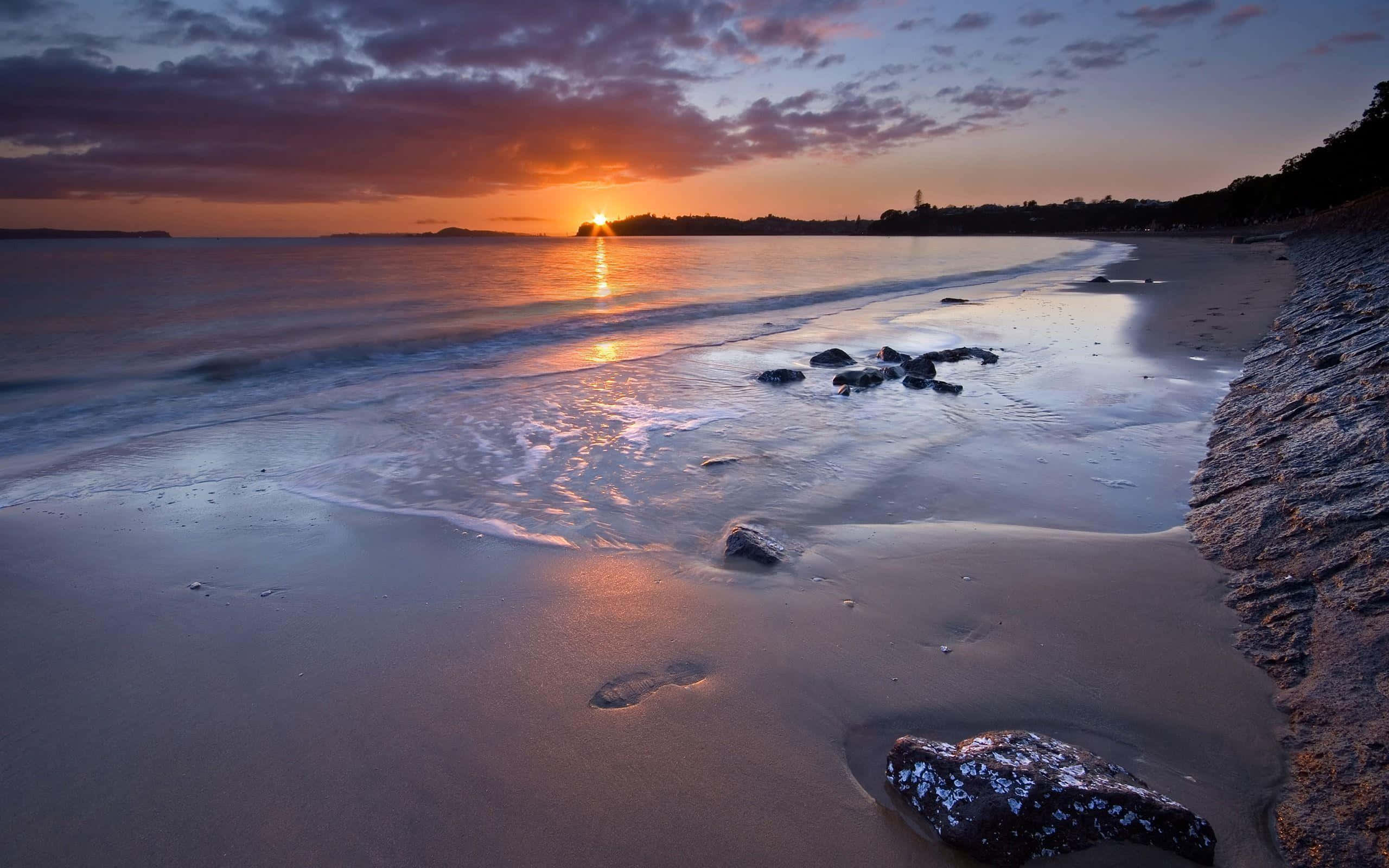 Beach At Night 桌布 壁紙 — 夕陽將天空染成火橙色和紫色，溫柔的海浪輕拍著佈滿岩石和腳印的沙灘。