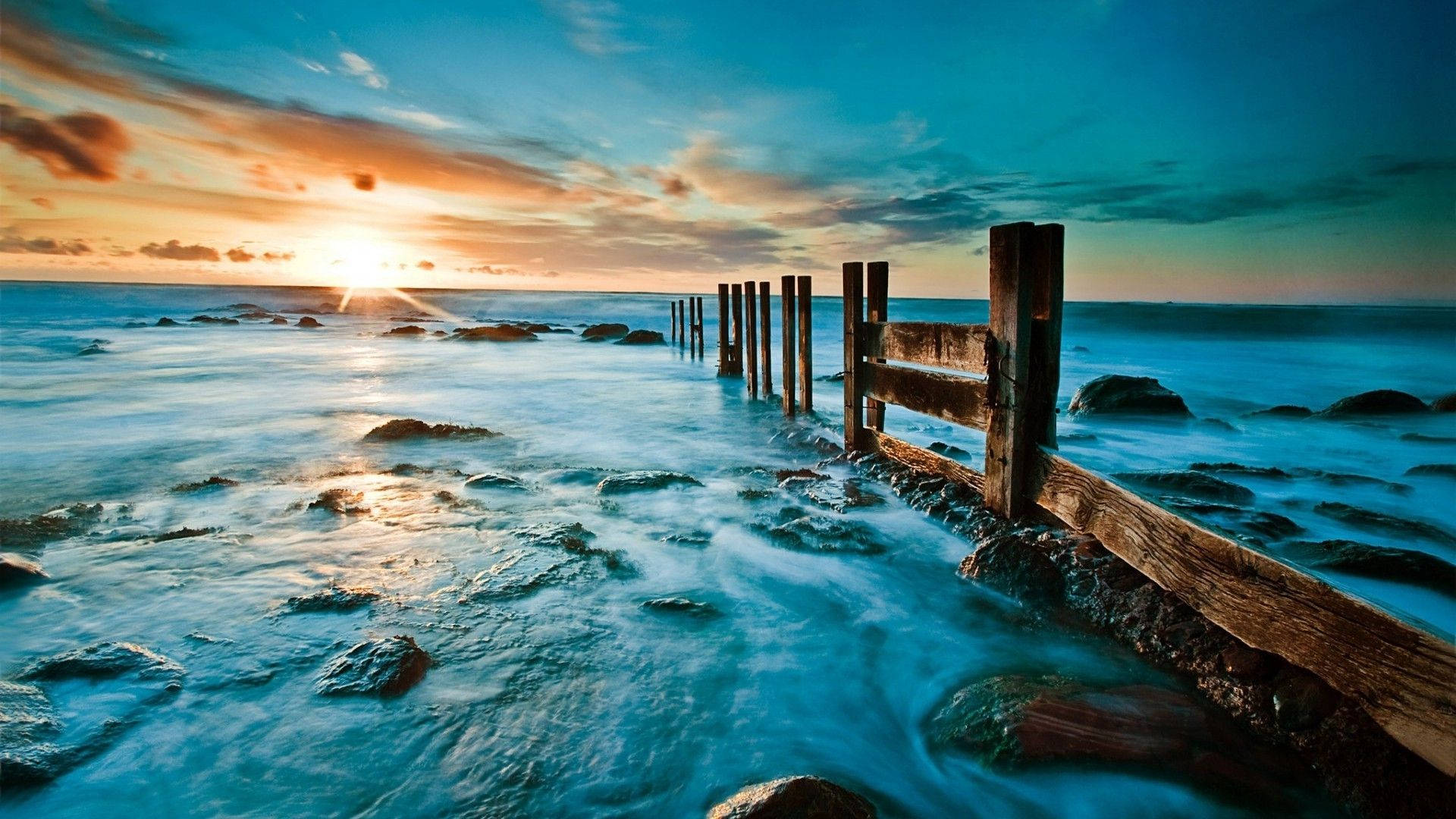 View 桌布 壁紙 — 夕陽將天空染成火橙色和藍綠色，海浪在海洋邊緣風化的木樁周圍流動。