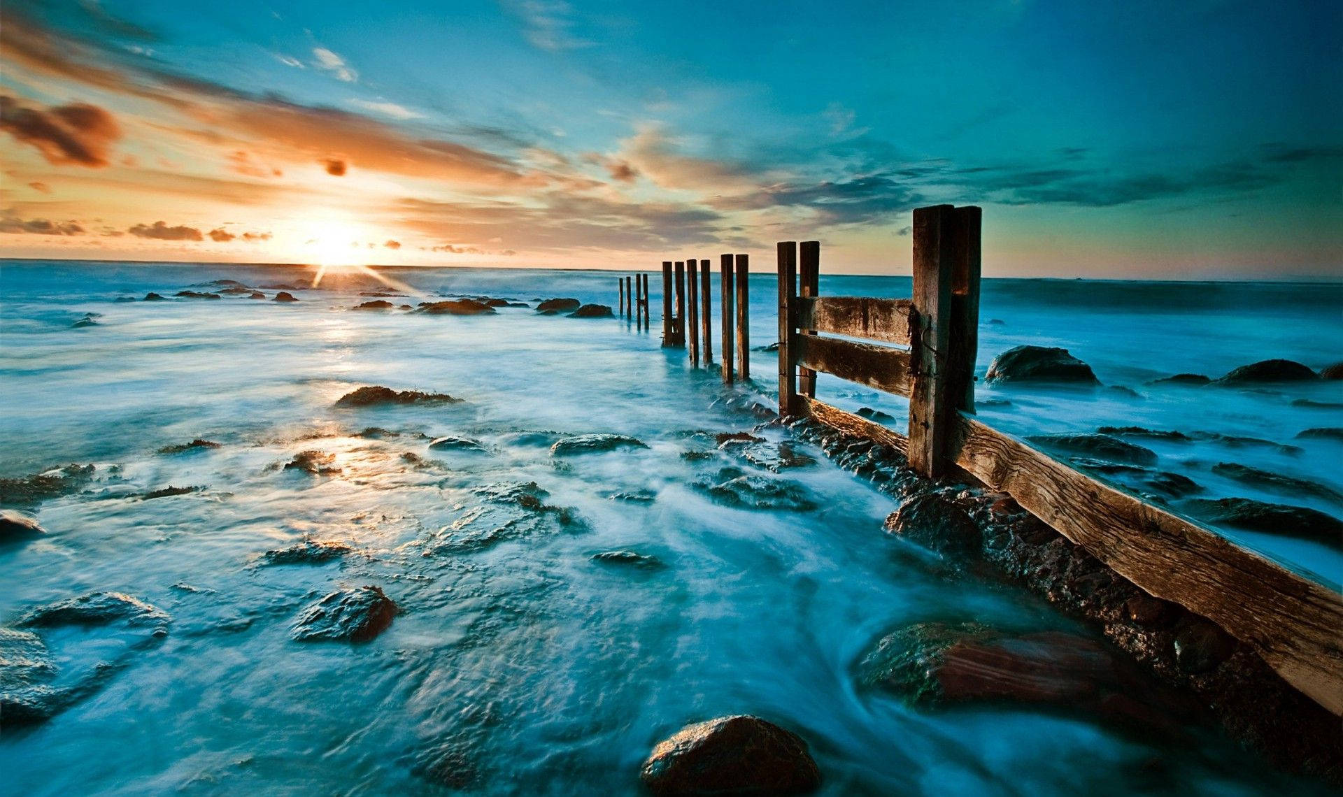 綠色天空 桌布 壁紙 — 夕陽將天空染成火橙色和青綠色，海浪平穩地流過風化的木樁和岩石。