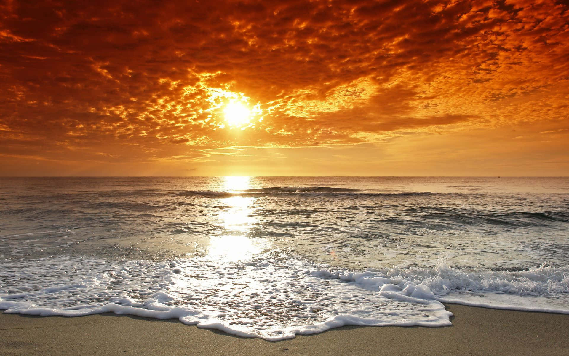 海灘日落 桌布 壁紙 — 夕陽將天空染成火熱的橙色，海浪拍打沙灘，金色光芒在水面上反射。
