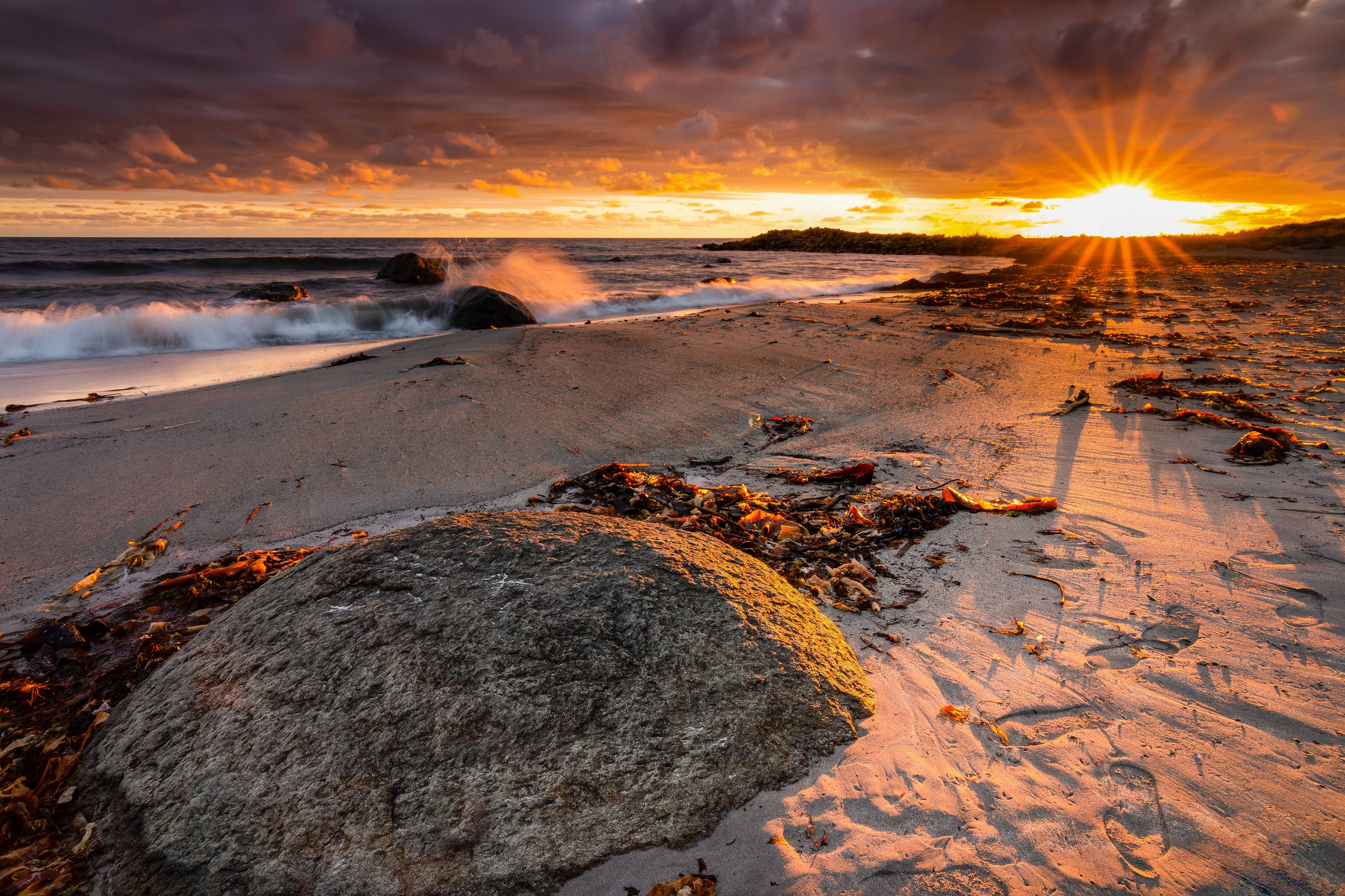 Norway 桌布 壁紙 — 夕陽將天空染成火熱的橙色，海浪拍打著岩石，沙灘上散落著海藻。