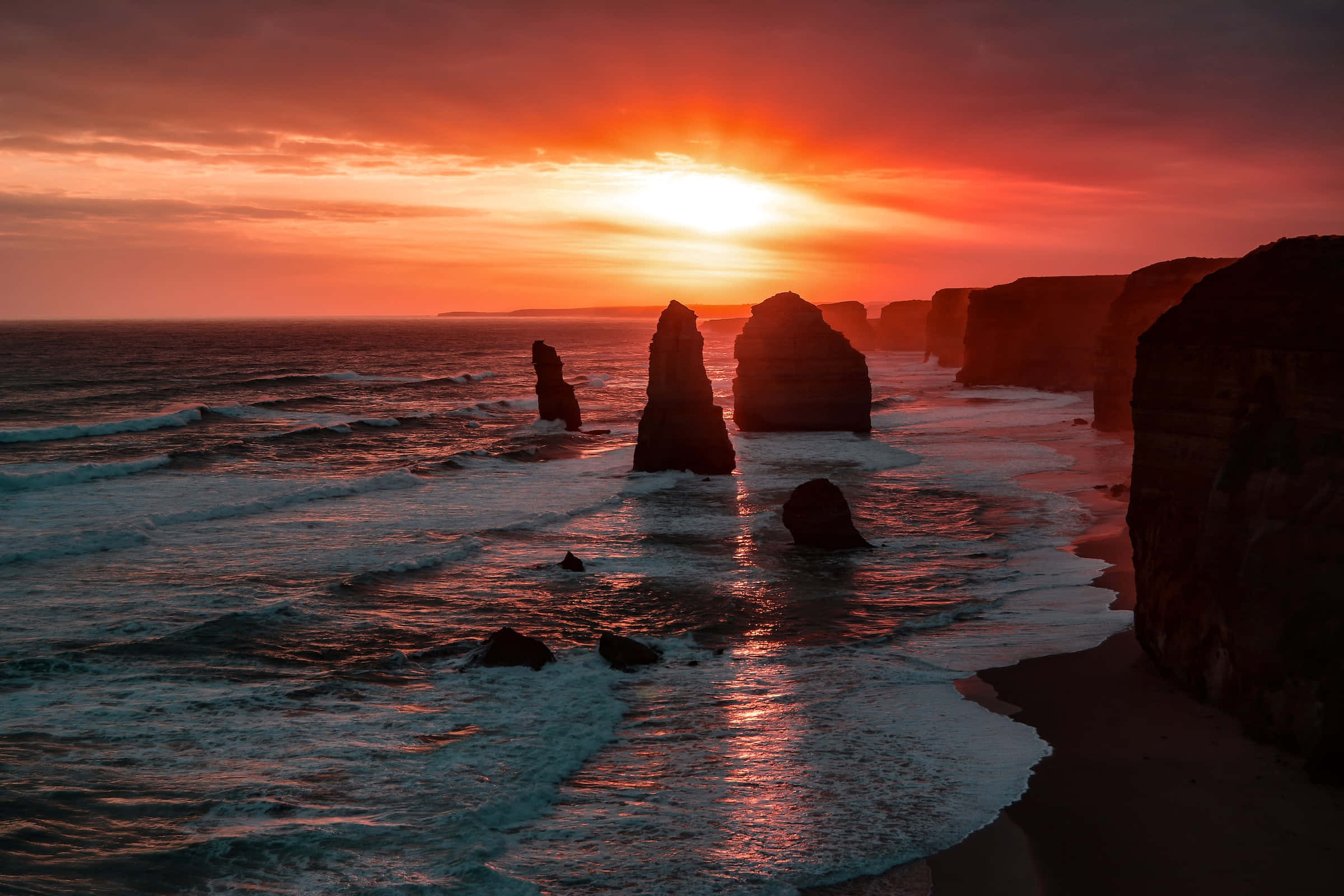 Breathtaking 桌布 壁紙 — 夕陽將天空染成火熱的橙色，海浪拍打著崎嶇海岸線上高聳的石灰岩柱。