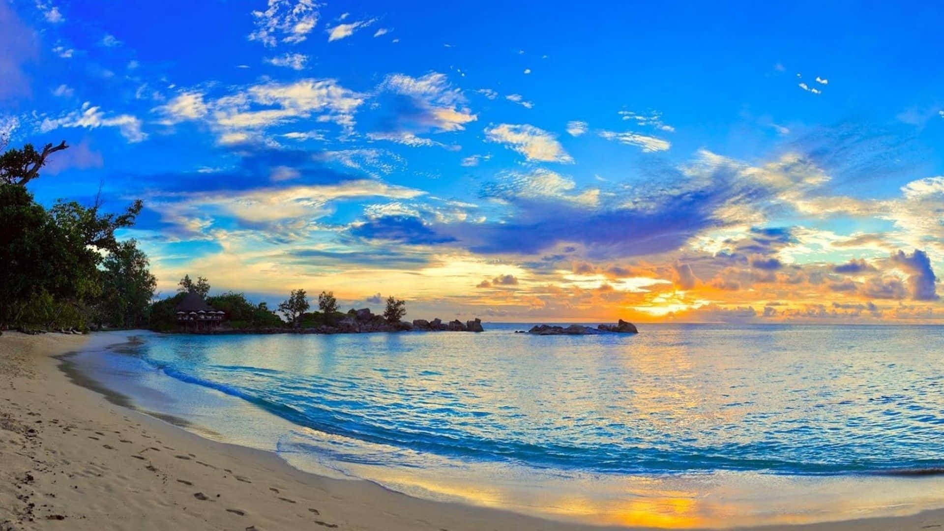 全高清 桌布 壁紙 — 夕陽將天空染成火熱的橙色和深藍色，海浪輕輕拍打著純淨的白色沙灘，沙灘兩旁綠樹成蔭。