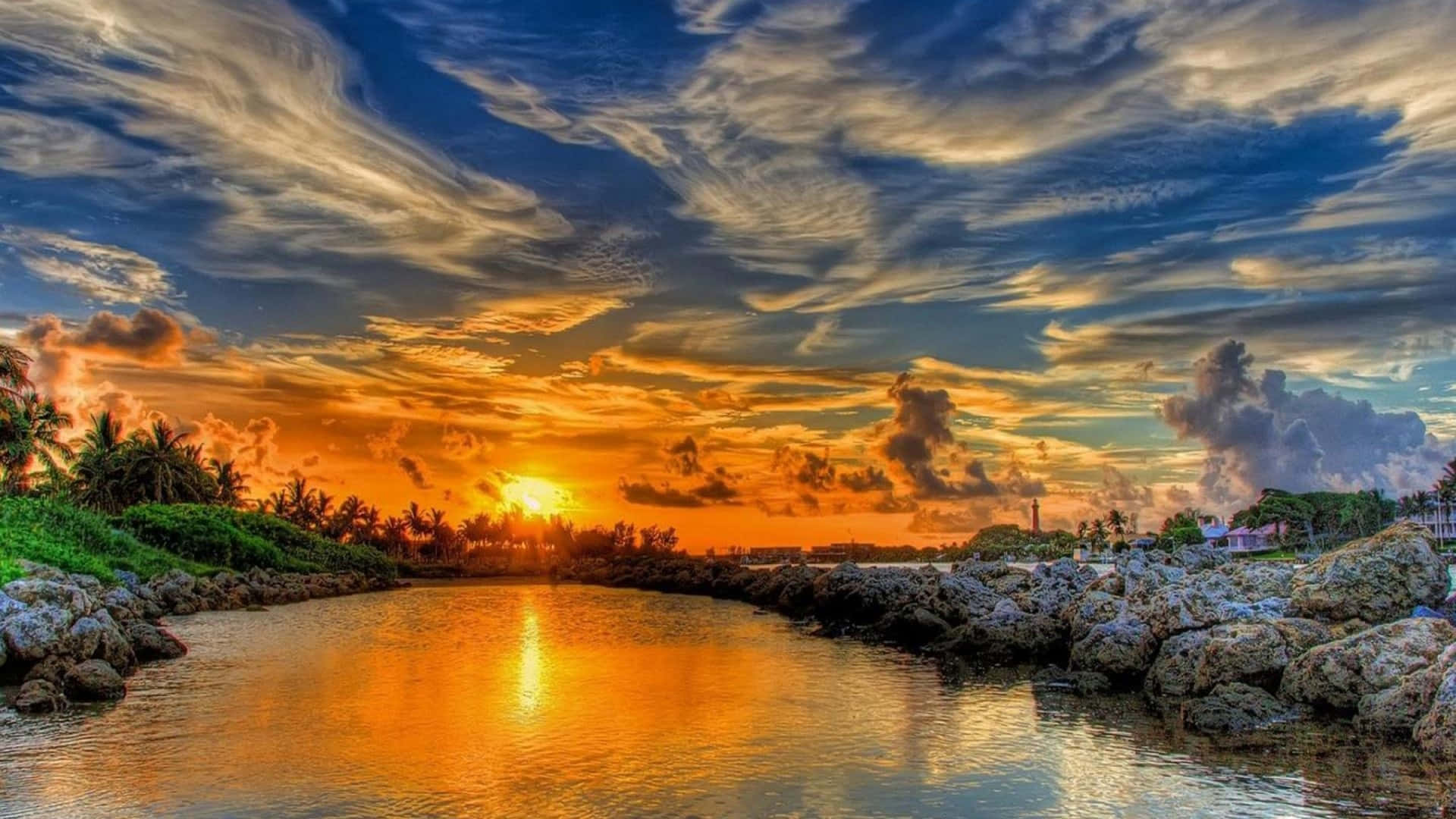 Glorious 桌布 壁紙 — 夕陽將天空染成火熱的橙色和深藍色，倒映在平靜的水面上，四周環繞著岩石海岸和棕櫚樹。