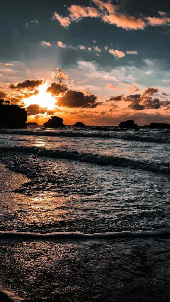 Live Aesthetic 桌布 壁紙 — 夕陽將天空染成火熱的橙色和粉紅色，海浪拍打著寧靜海灘上的黑色岩石。