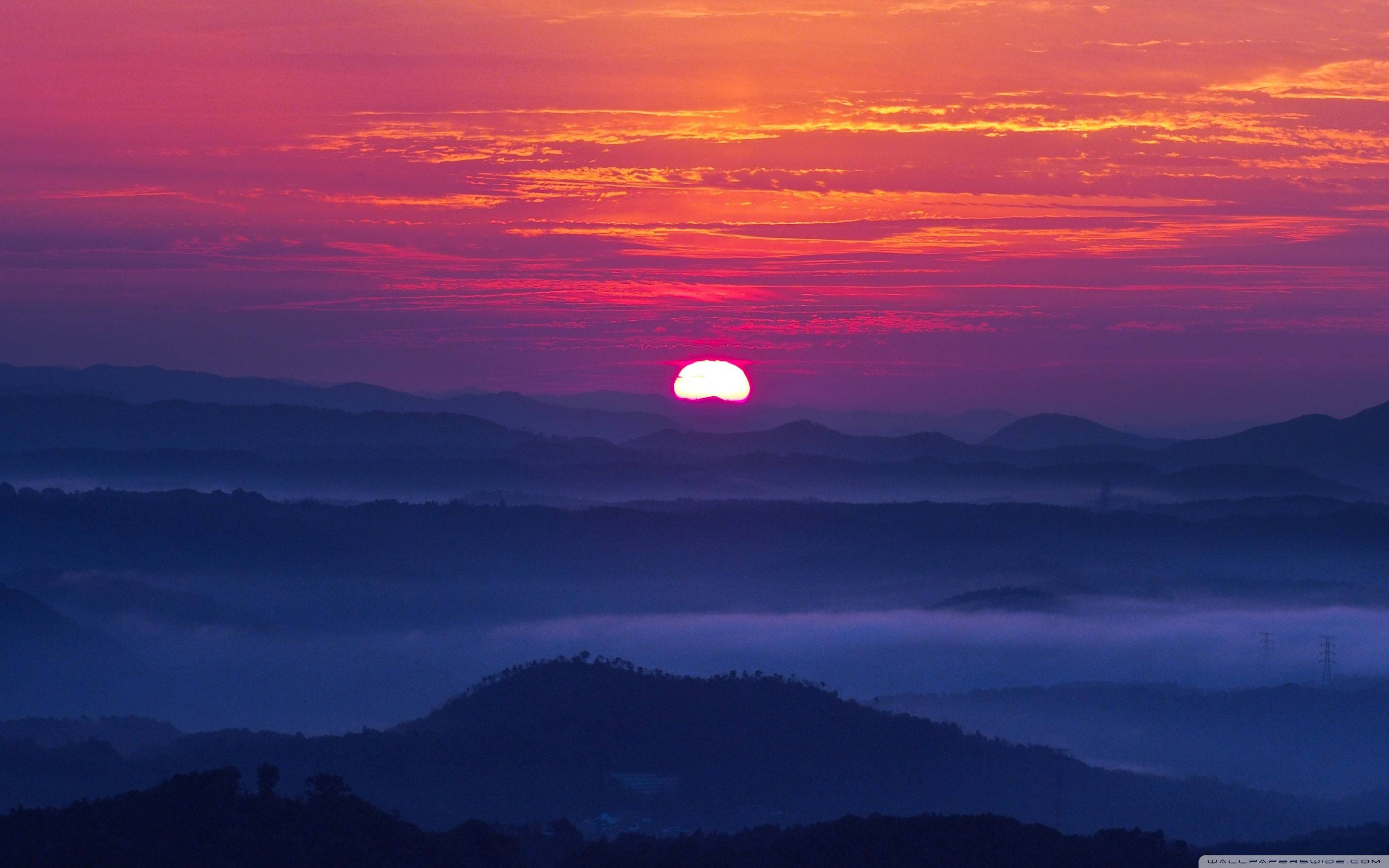 日落 桌布 壁紙 — 夕陽將天空染成火熱的橙色和粉紅色，太陽沉入層層疊疊、霧氣繚繞的山脊之後。
