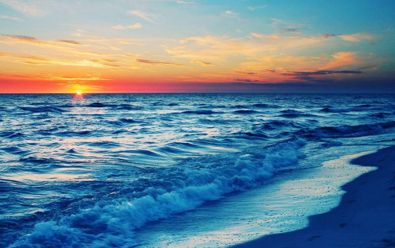 海灘日落 桌布 壁紙 — 夕陽將天空染成火熱的橙色和粉紅色，海浪在廣闊的海洋下拍打著沙灘。