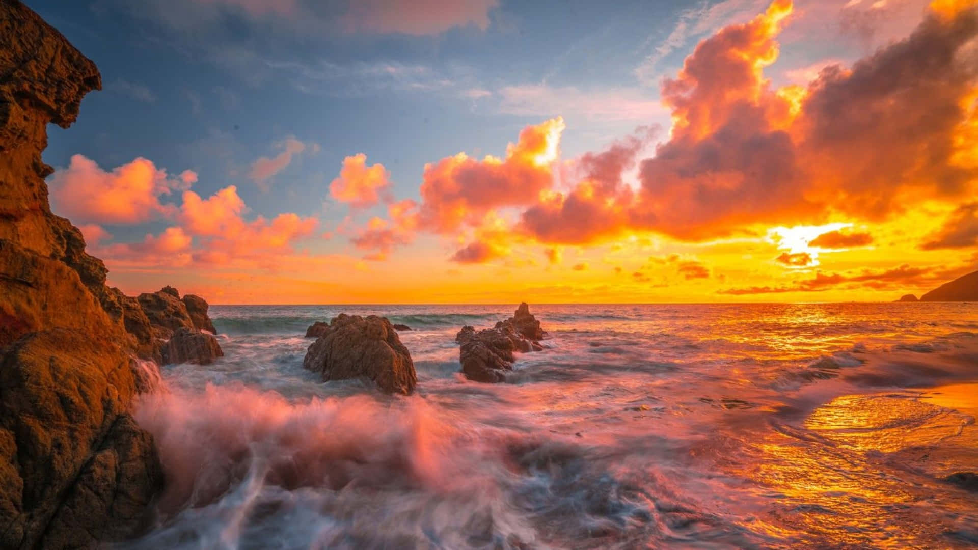 海灘 桌布 壁紙 — 夕陽將天空染成火熱的橙色和粉紅色，海浪拍打著崎嶇的海岸岩石。