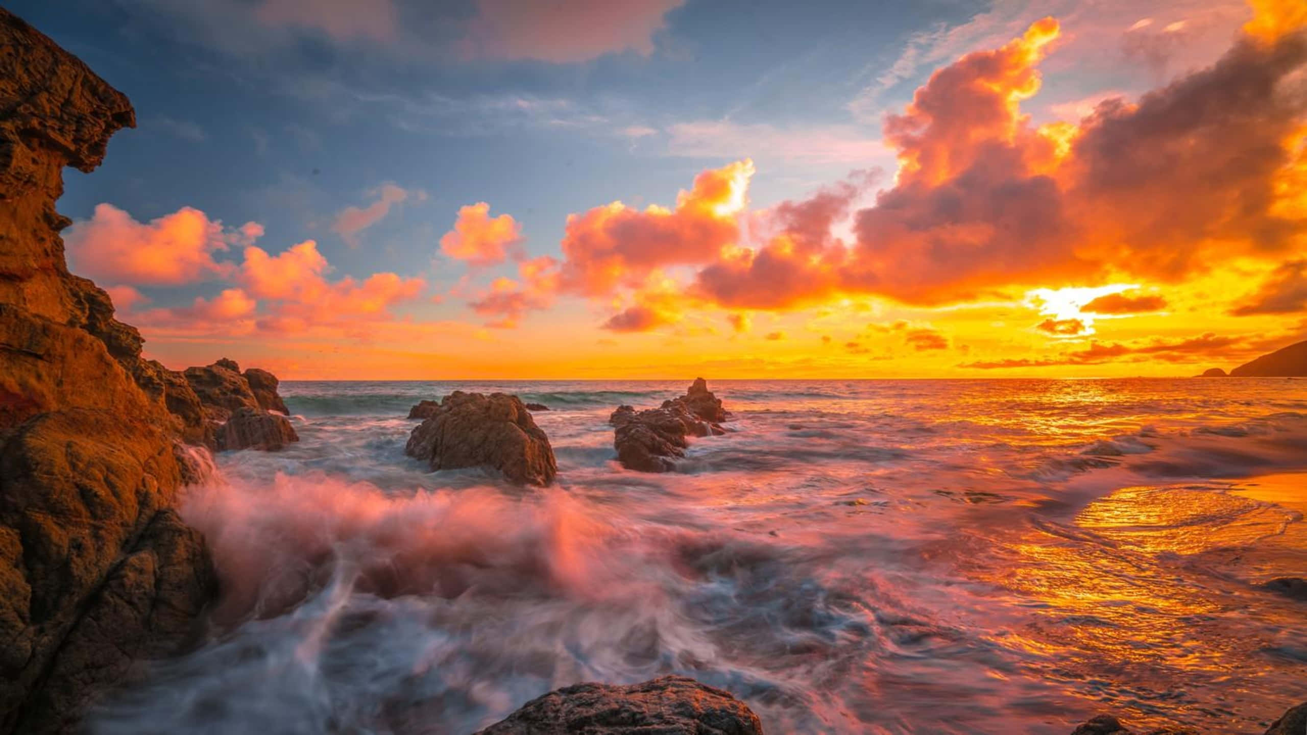 Ocean Sunset 桌布 壁紙 — 夕陽將天空染成火熱的橙色和粉紅色，海浪拍打著崎嶇的海岸岩石。