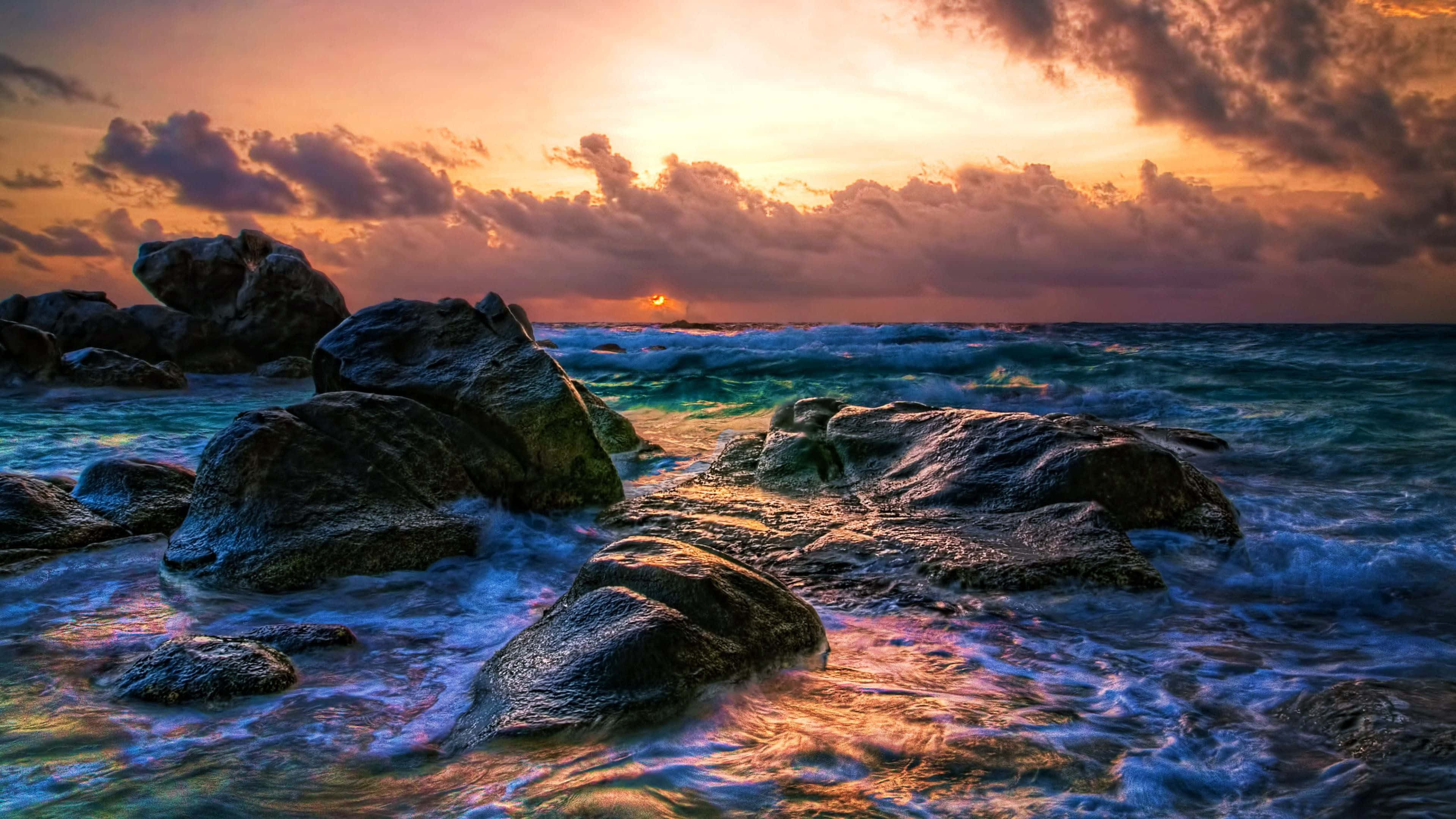 極高畫質 桌布 壁紙 — 夕陽將天空染成火熱的橙色和紫色，海浪拍打著海岸的黑色岩石。