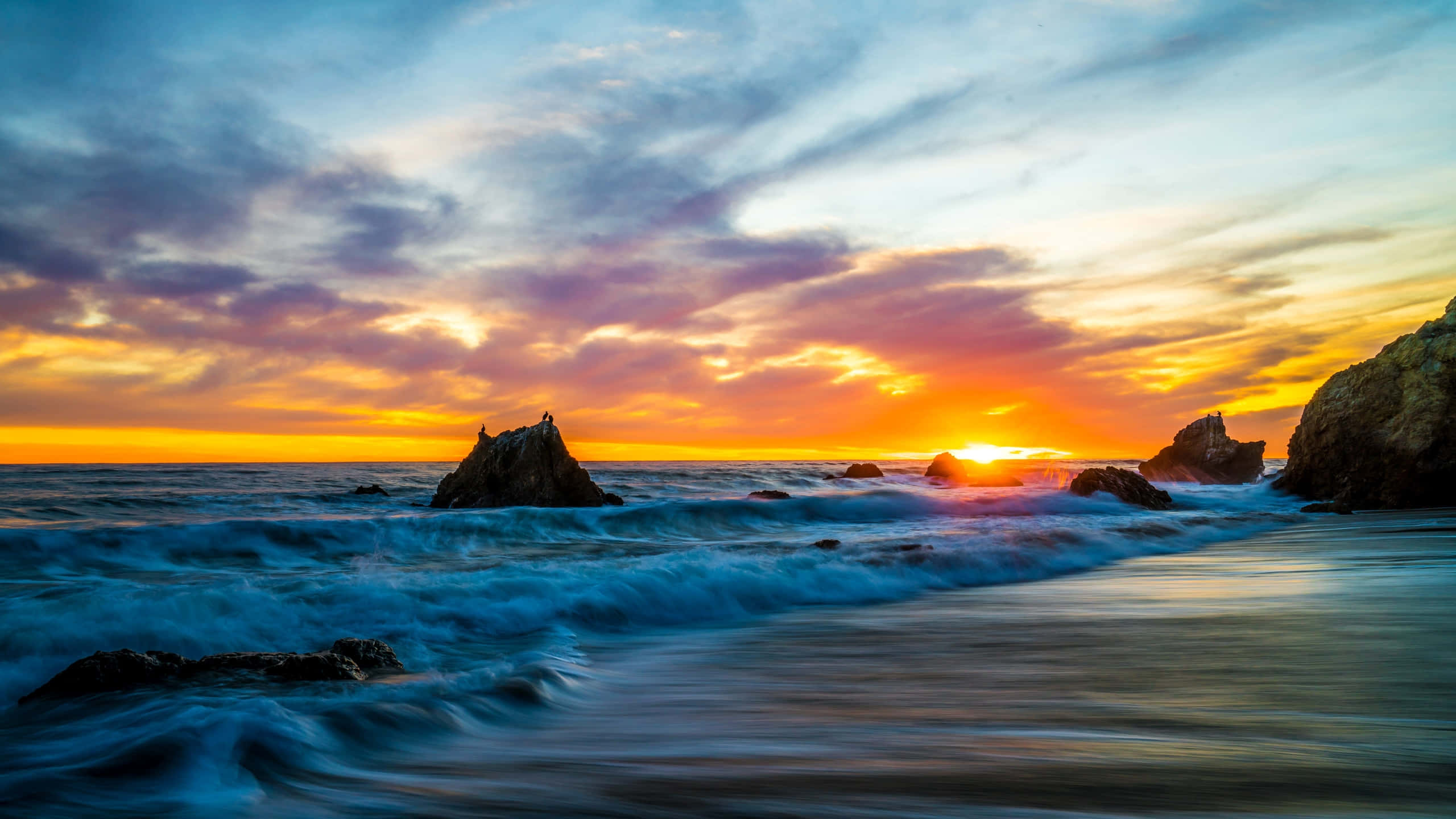 1440p Malibu 桌布 壁紙 — 夕陽將天空染成火熱的橙色和紫色，海浪拍打著崎嶇的海岸岩石。